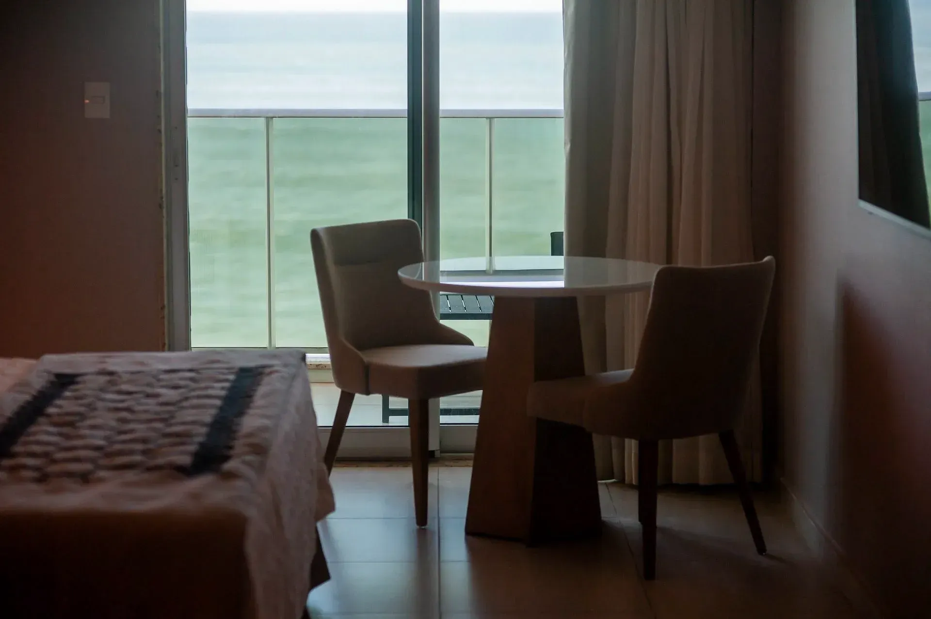 Quarto de hotel com duas cadeiras e uma mesa redonda junto à janela com vista para o oceano.