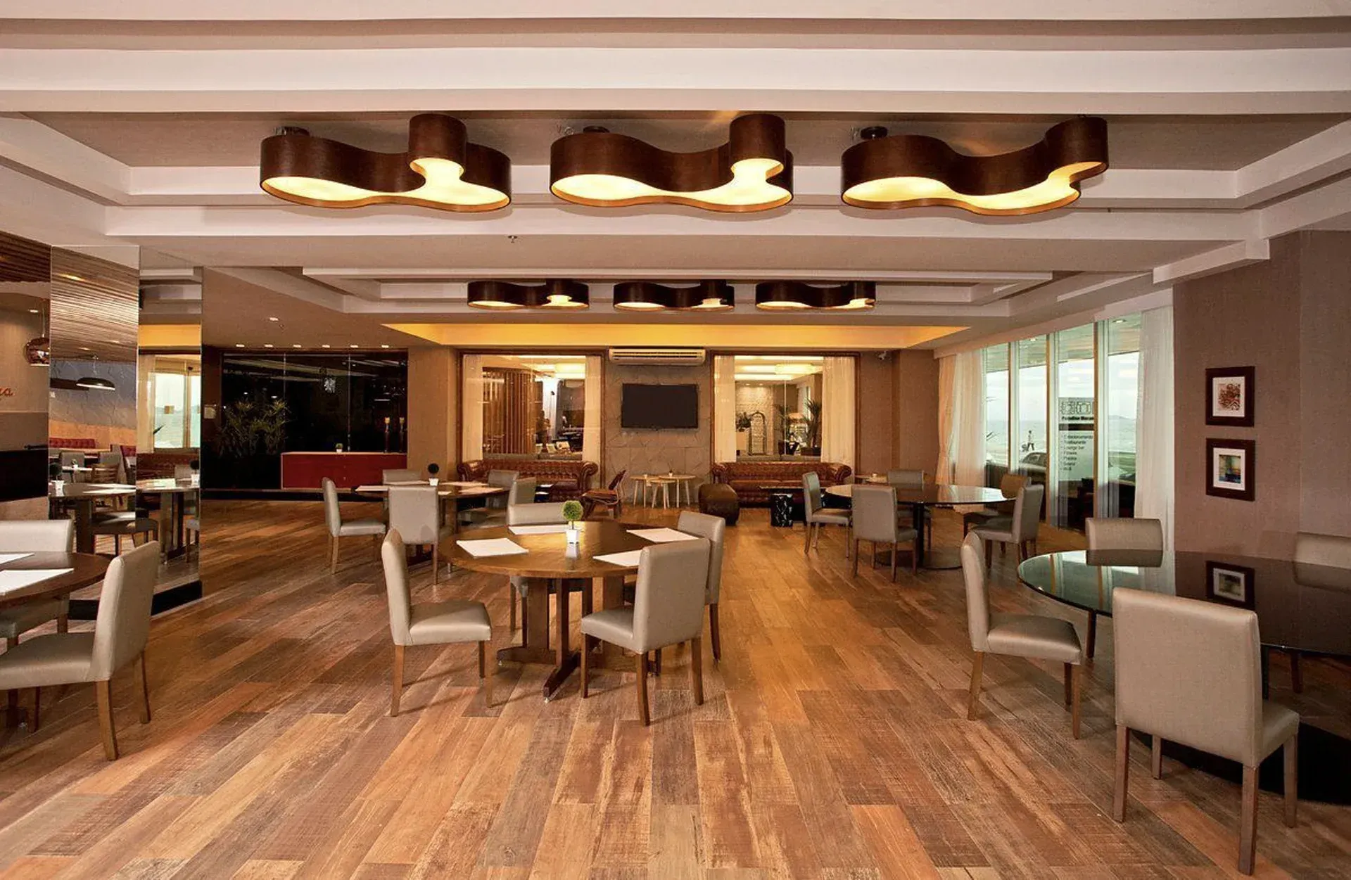 Sala de estar moderna do hotel com piso de madeira, mesas, cadeiras e iluminação pendente aconchegante.