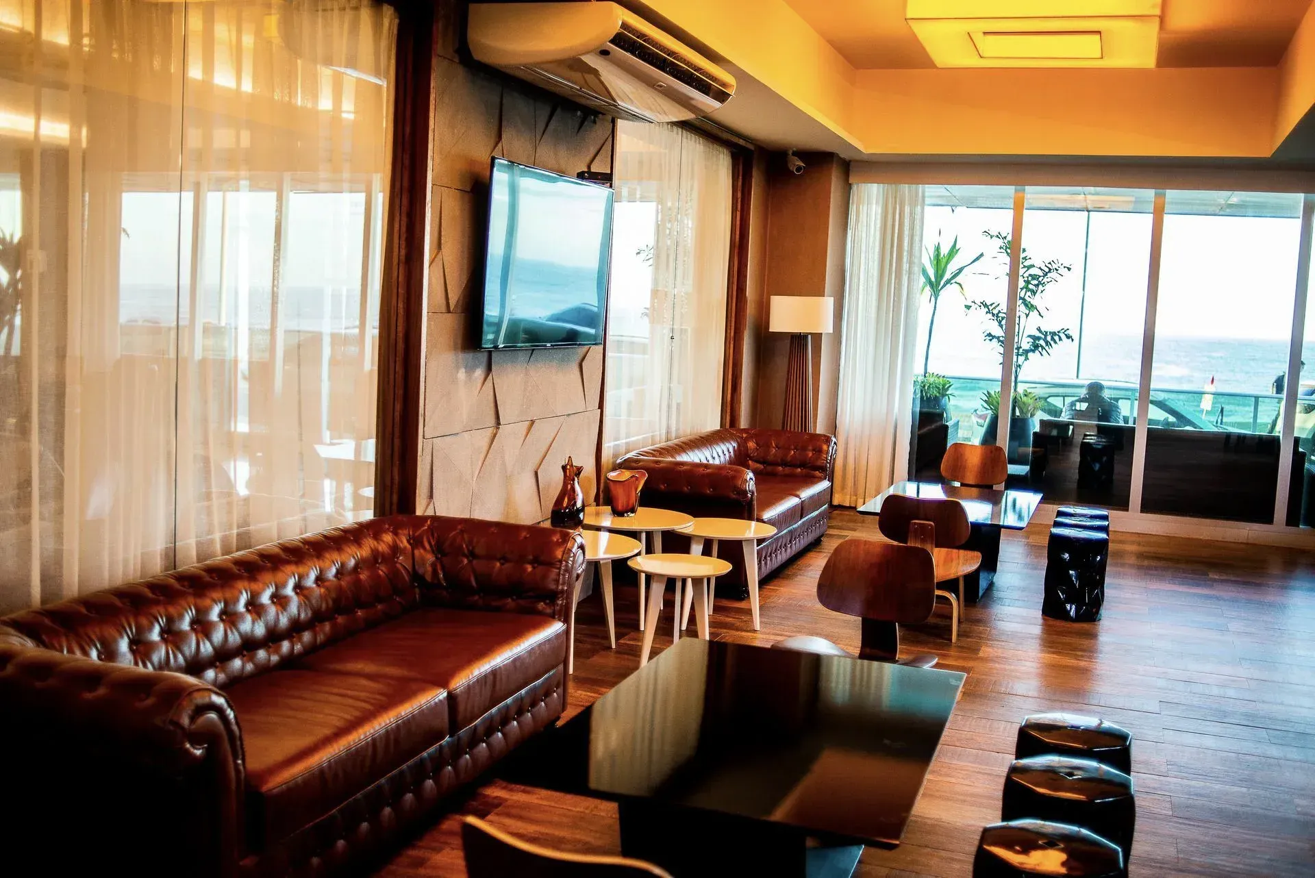 Sala de estar moderna com sofás de couro, piso de madeira, TV de parede e grandes janelas com vista para uma varanda.