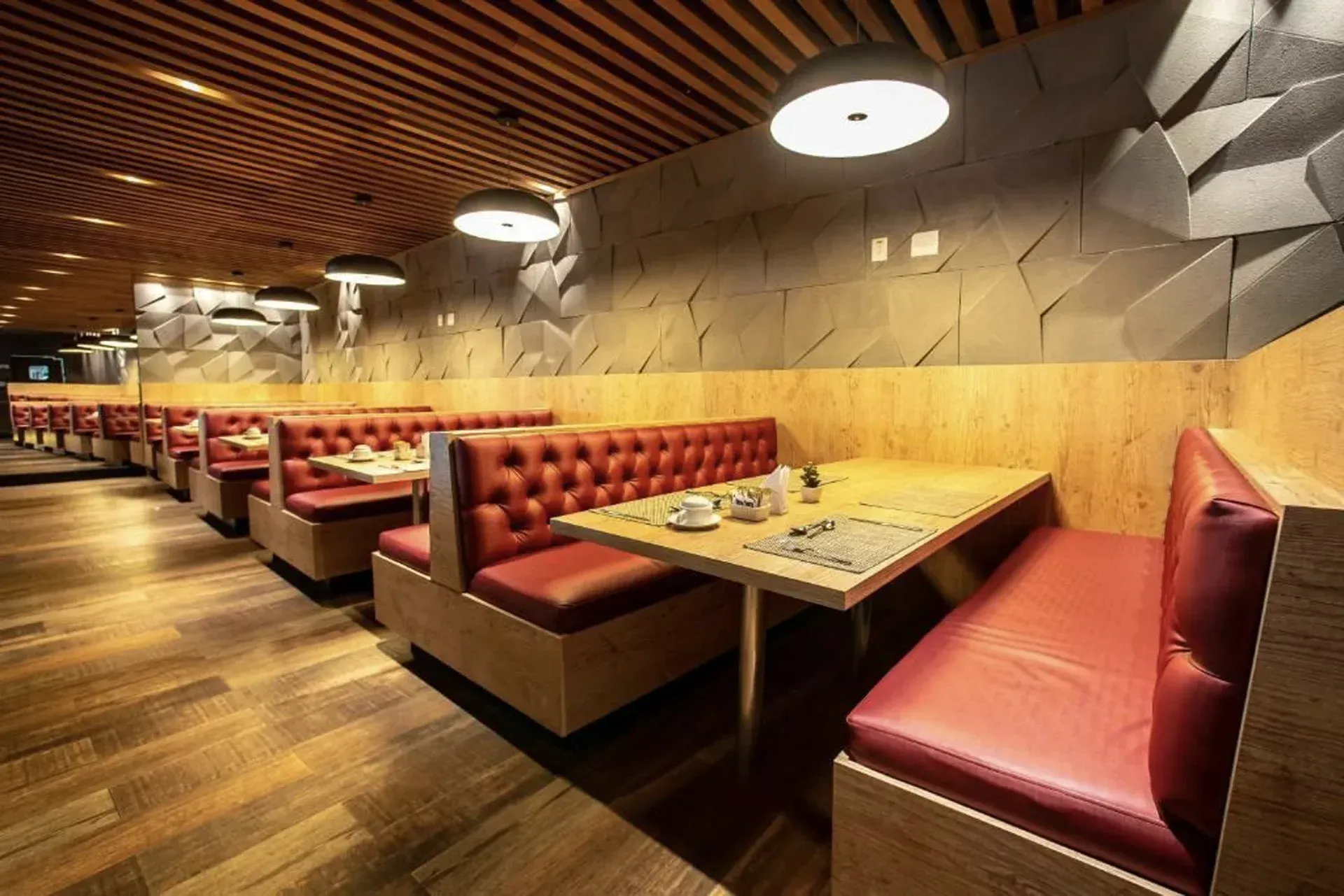 Cabines de restaurante vazias com bancos de vinil vermelho, mesas de madeira e decoração geométrica na parede.