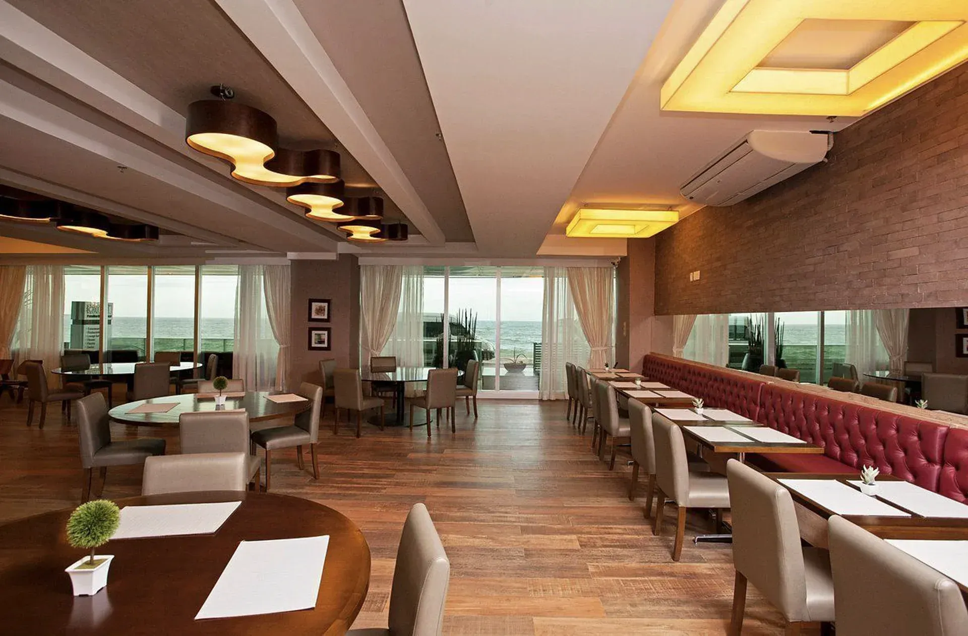 Interior vazio de um restaurante moderno com piso de madeira, cadeiras bege, bancos vermelhos e grandes janelas com vista.