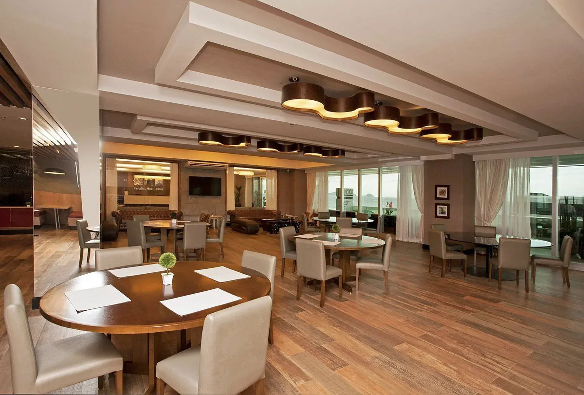 Sala de jantar moderna em plano aberto com piso de madeira, cadeiras brancas e grandes janelas com vista para a vegetação.