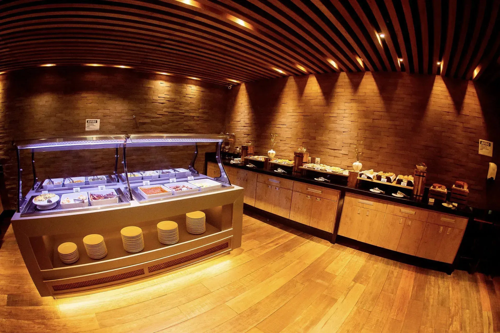 Restaurante buffet com paredes de tijolos e iluminação suave, balcão de saladas de vidro e teto de madeira aconchegante.