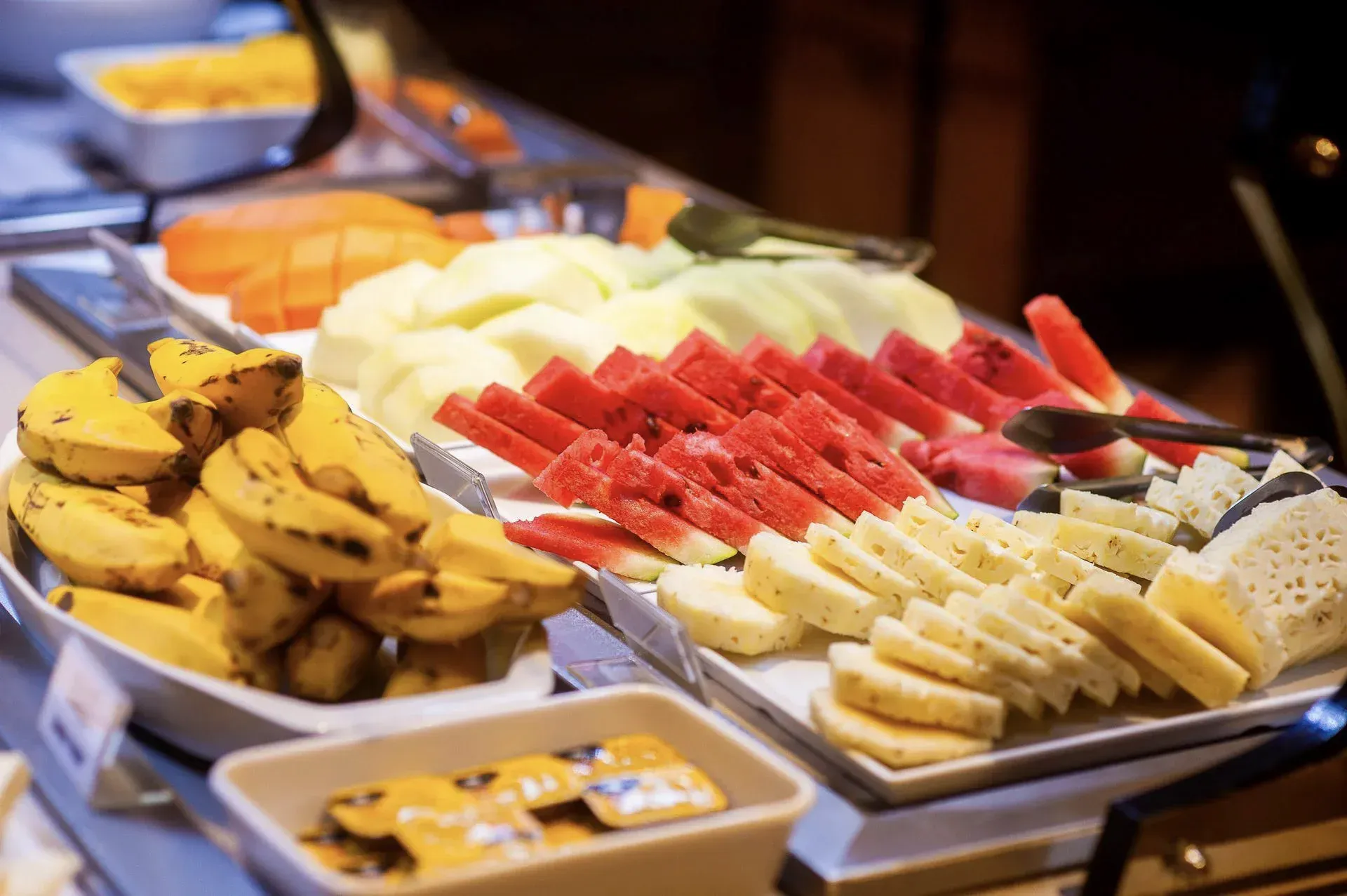 Prato de frutas com bananas, abacaxi, melancia e melão dispostos em bandejas de buffet.