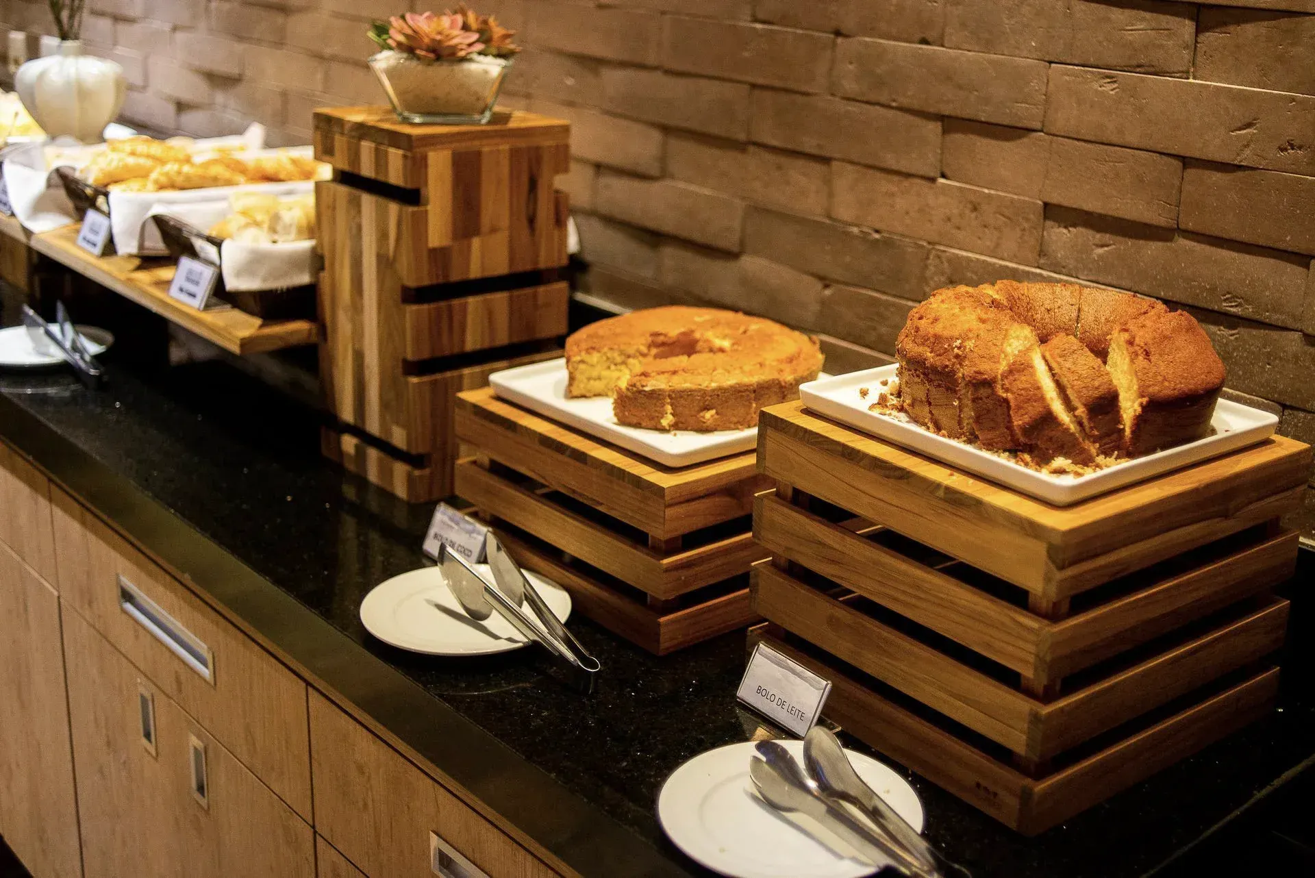 Mesa de buffet com bandejas de madeira empilhadas contendo pães e doces encostada em uma parede de tijolos.