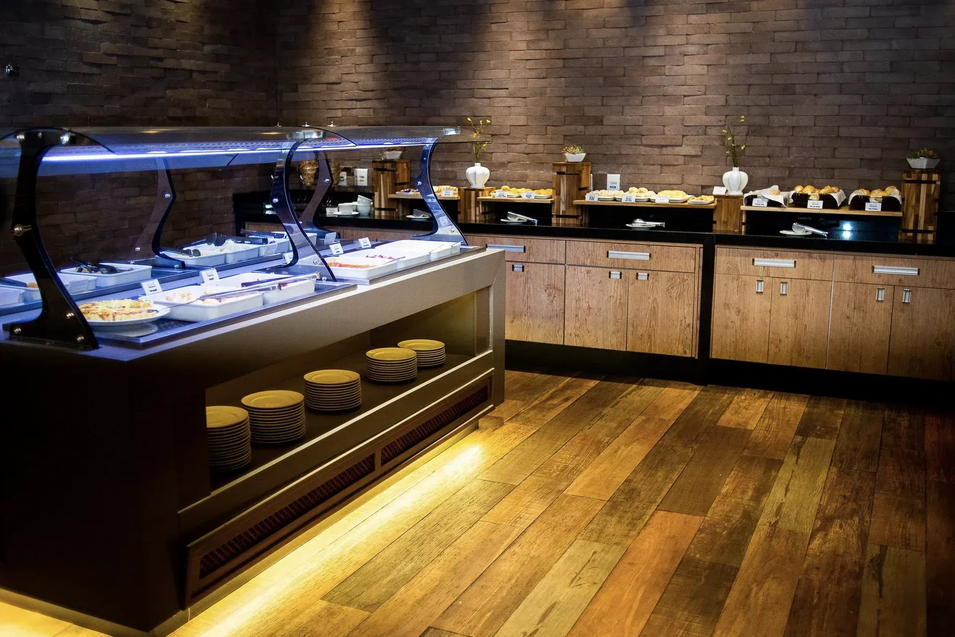 Balcão de buffet vazio com travessas cobertas, piso de madeira e parede de tijolos em uma área de refeições de restaurante.
