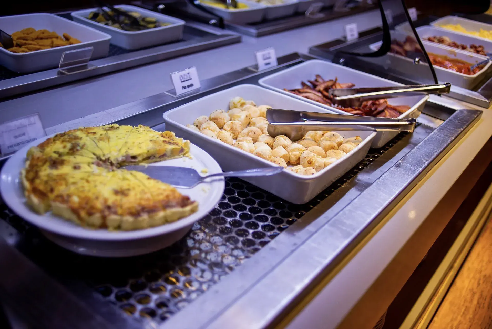 Balcão de buffet com bandejas de comida quente variada, incluindo assados ​​e bolinhos, sob luzes aquecedoras.