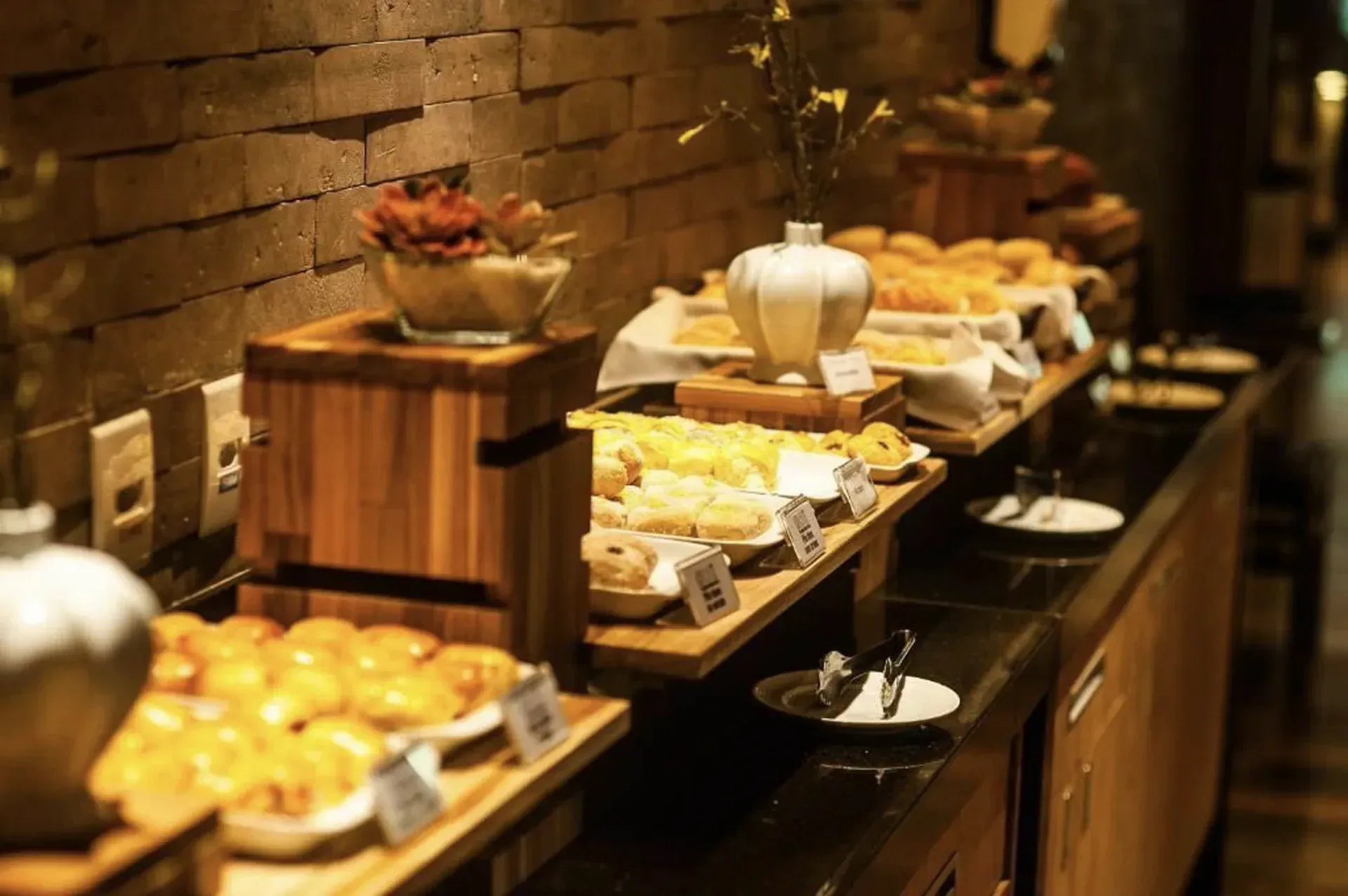 Buffet de padaria com doces, pães, frutas e chás em bandejas de vários andares ao longo de um balcão de madeira.