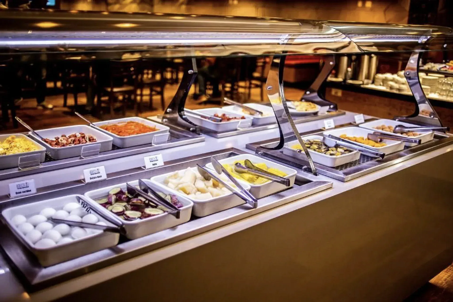 Apresentação de comida em estilo buffet com pratos variados em bandejas de metal sob iluminação aconchegante.