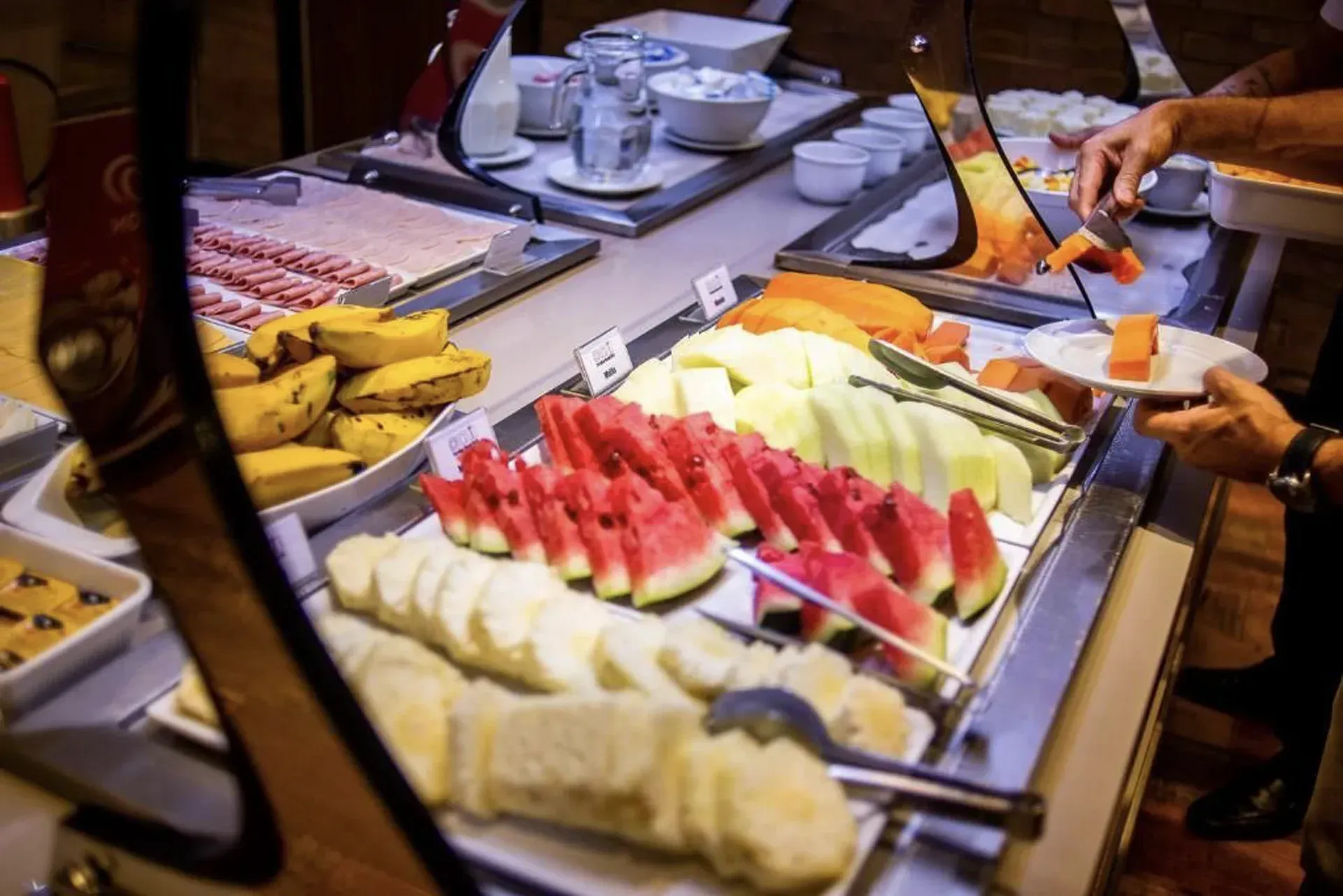 Prato de frutas com fatias de melancia e melão em uma mesa de bufê, com uma mão servindo a comida.