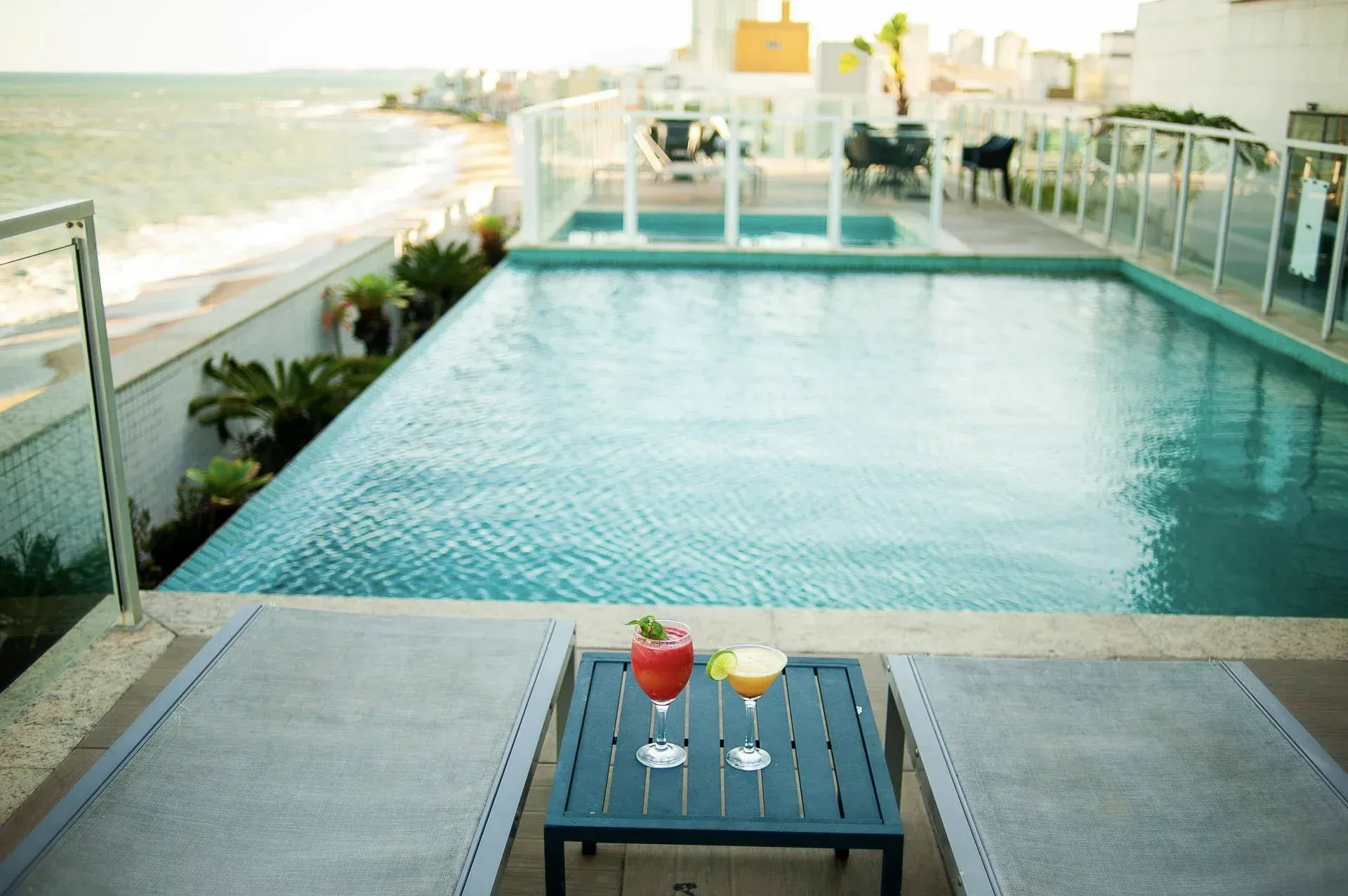 Piscina infinita na cobertura, à beira-mar, com uma pequena mesa e duas bebidas em primeiro plano.