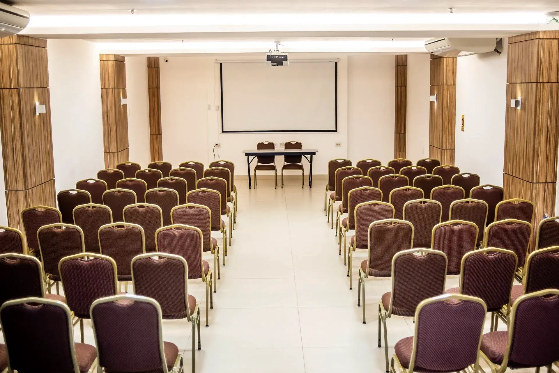 Sala de conferências vazia com fileiras de cadeiras marrons voltadas para uma tela de projeção e mesas na frente.