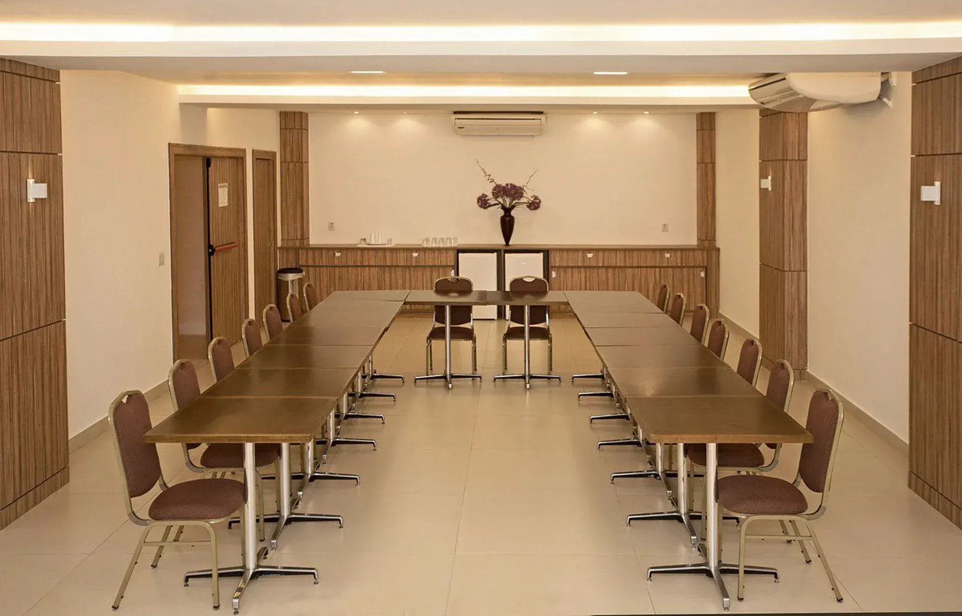Sala de conferências com mesas em formato de U, cadeiras marrons e um púlpito na frente, vista de trás.