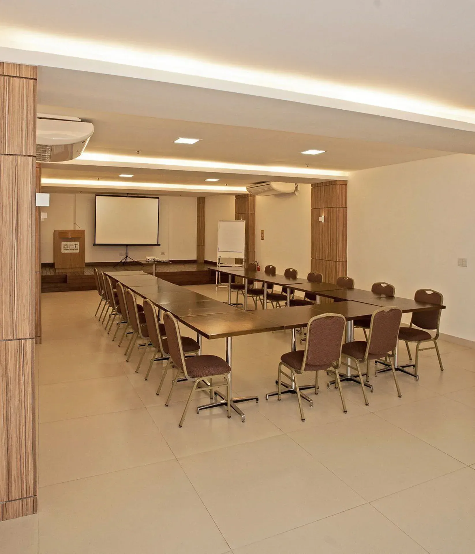 Sala de conferências vazia com mesas em formato de U, cadeiras, tela de projeção e luzes de teto.
