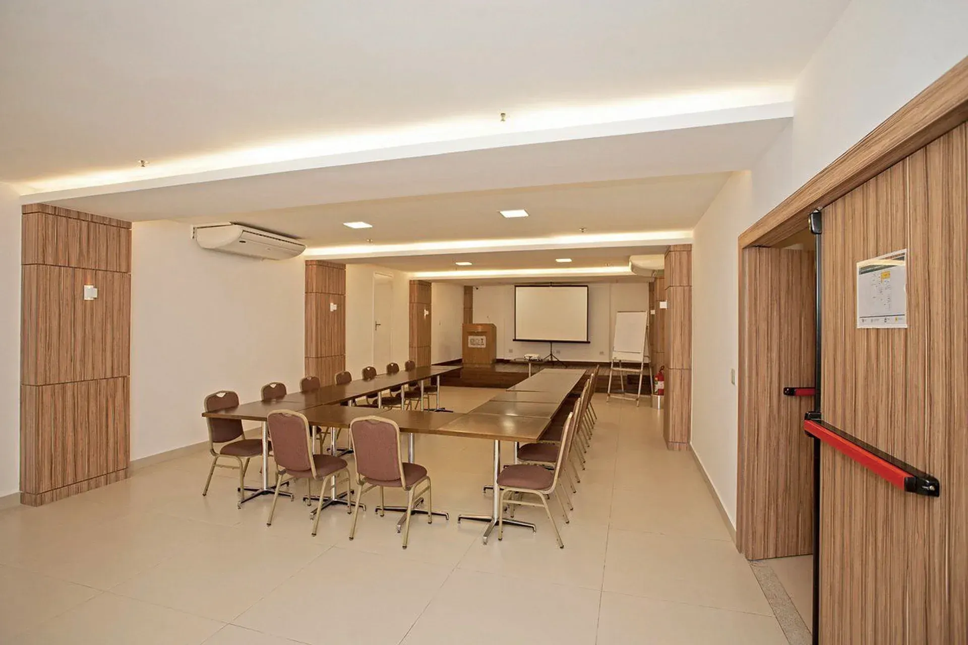Sala de conferências vazia com mesa comprida, cadeiras, tela de projeção e paredes revestidas de madeira.
