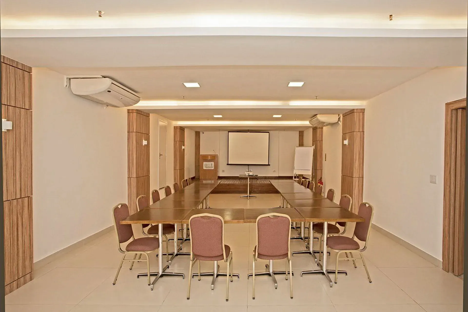 Sala de conferências com mesa comprida de madeira, cadeiras e tela de projeção.