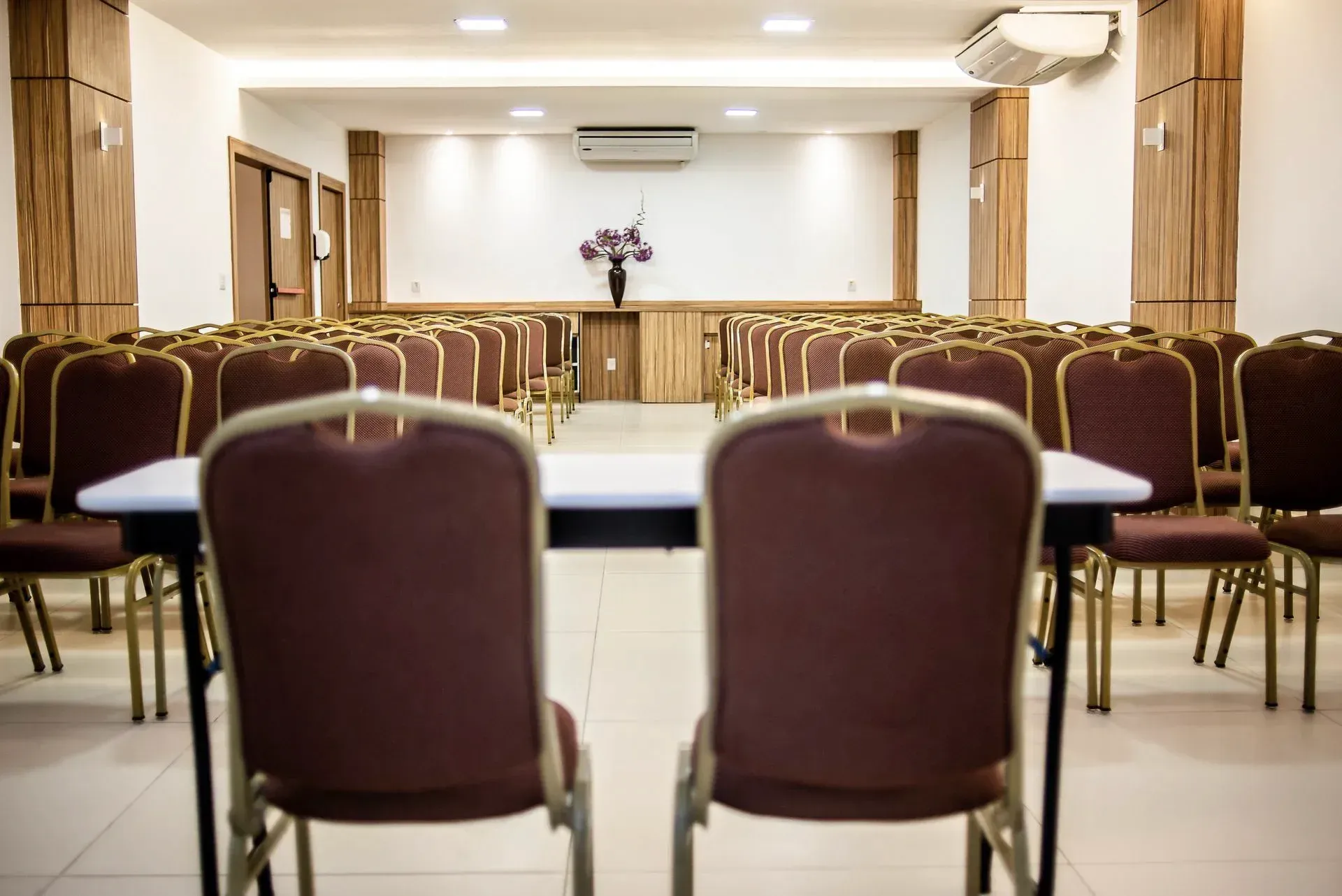 Sala de conferências vazia com fileiras de cadeiras cor de vinho e uma mesa à frente, iluminada por luzes de teto.