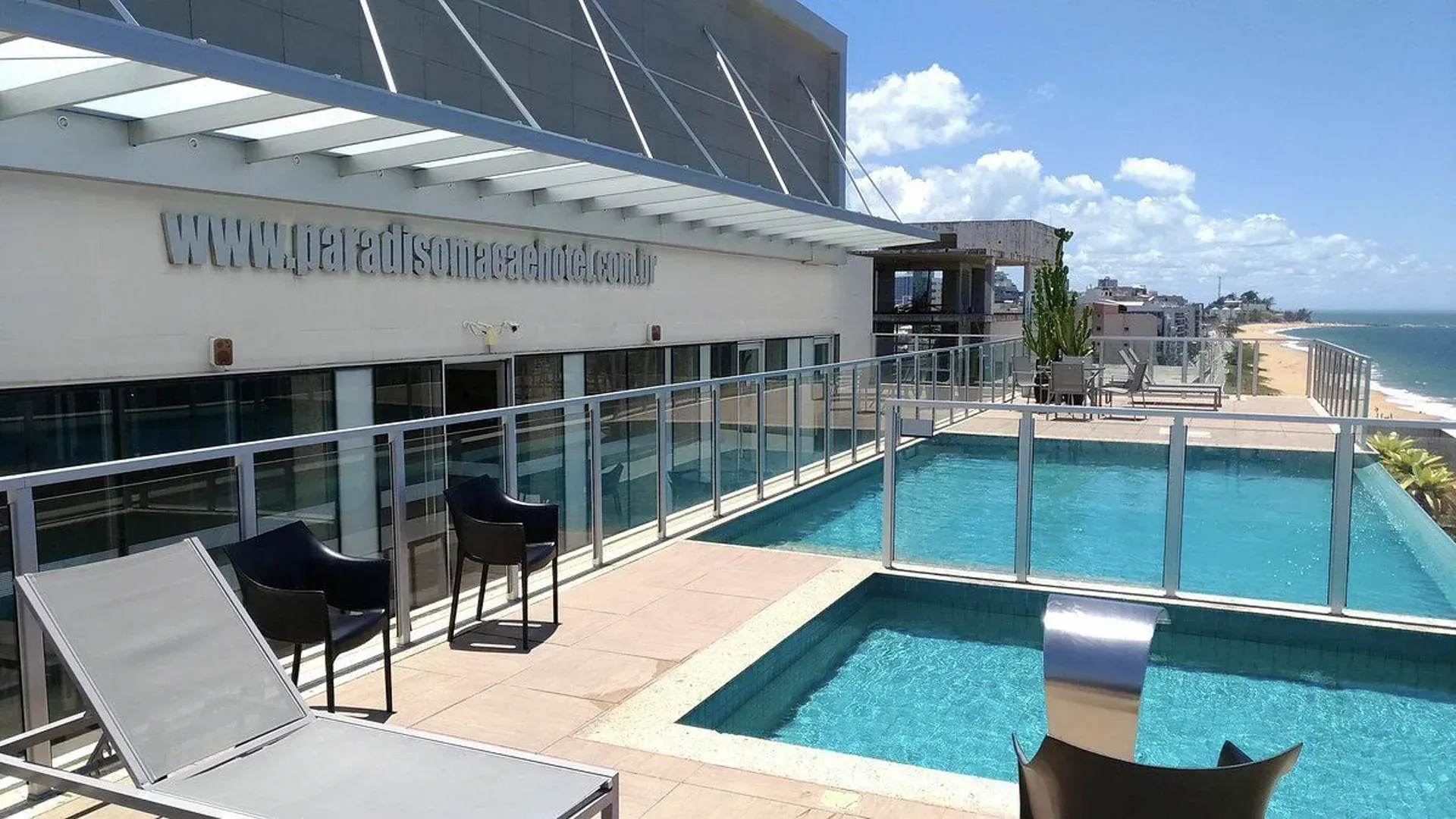 Terraço da piscina à beira-mar com espreguiçadeiras, guarda-corpo de vidro e vista para a praia sob um céu claro.