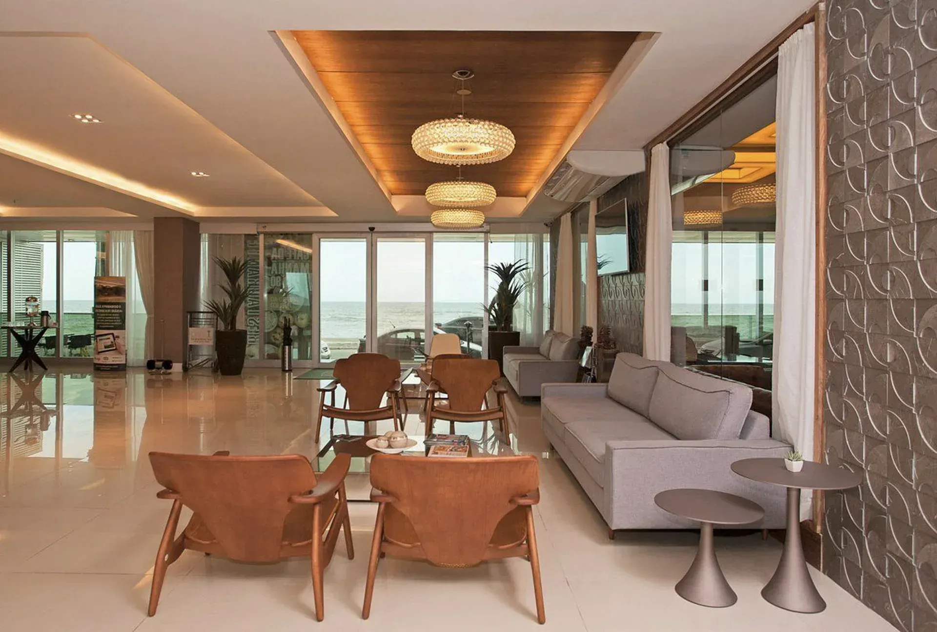 Sala de estar moderna com sofá cinza, cadeiras de madeira, paredes de vidro e iluminação aconchegante no teto, com vista.