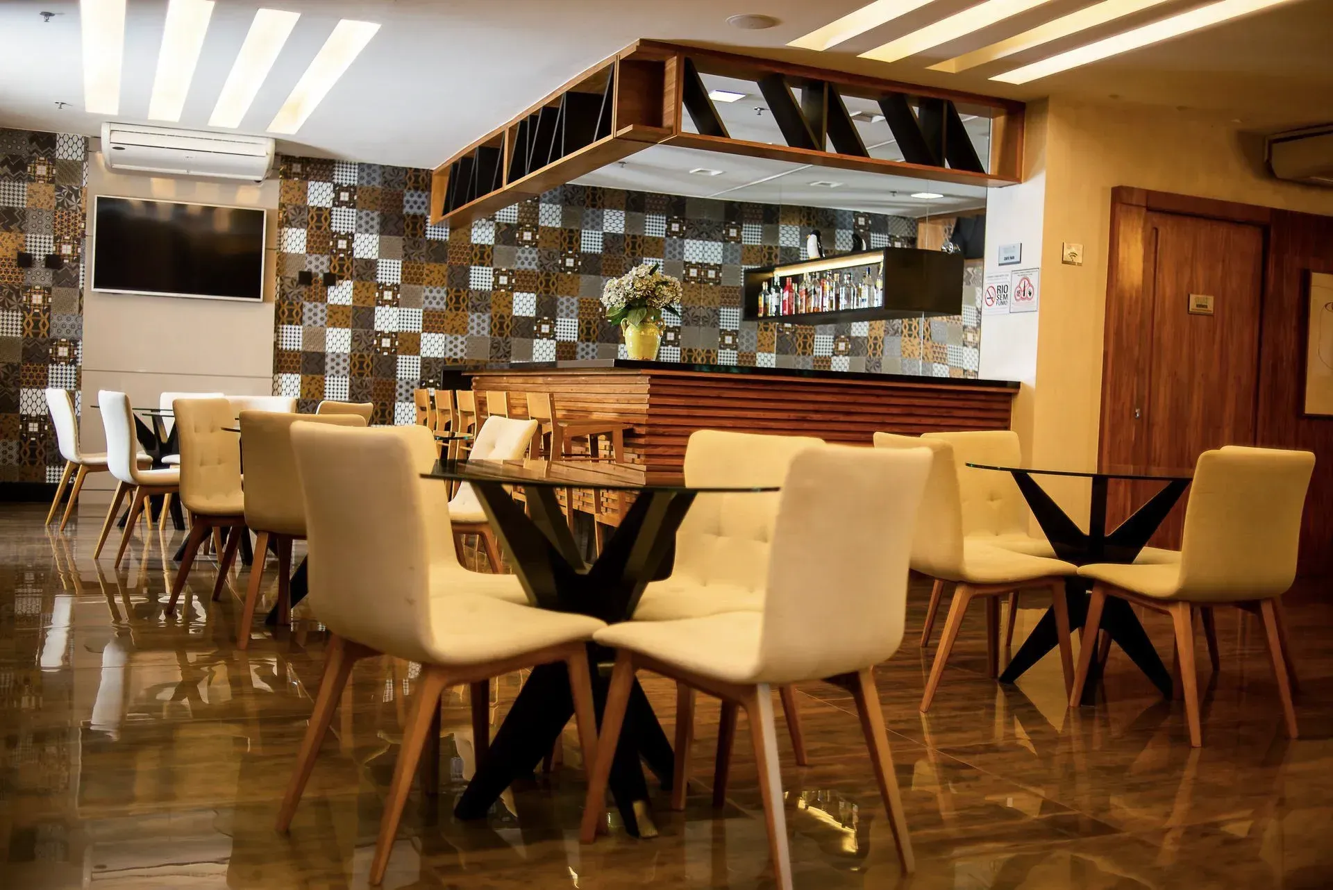Interior de restaurante moderno com mesas de madeira, cadeiras creme e área de bar espelhada.