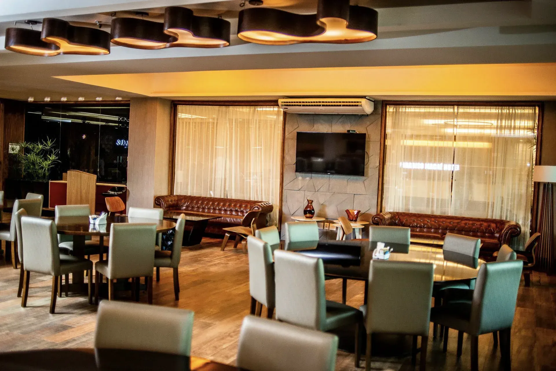 Sala de jantar moderna de restaurante com mesas de madeira, cadeiras brancas e azul-turquesa e iluminação.