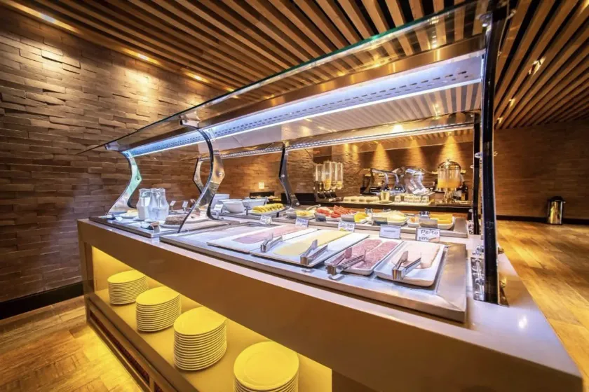 Balcão buffet com painéis de madeira, iluminação aconchegante e bandejas de comida atrás de vidro.