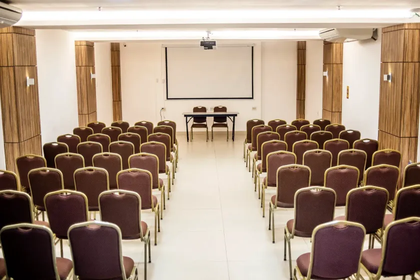 Sala de conferências vazia com fileiras de cadeiras roxas voltadas para uma tela de projeção e uma mesa à frente.