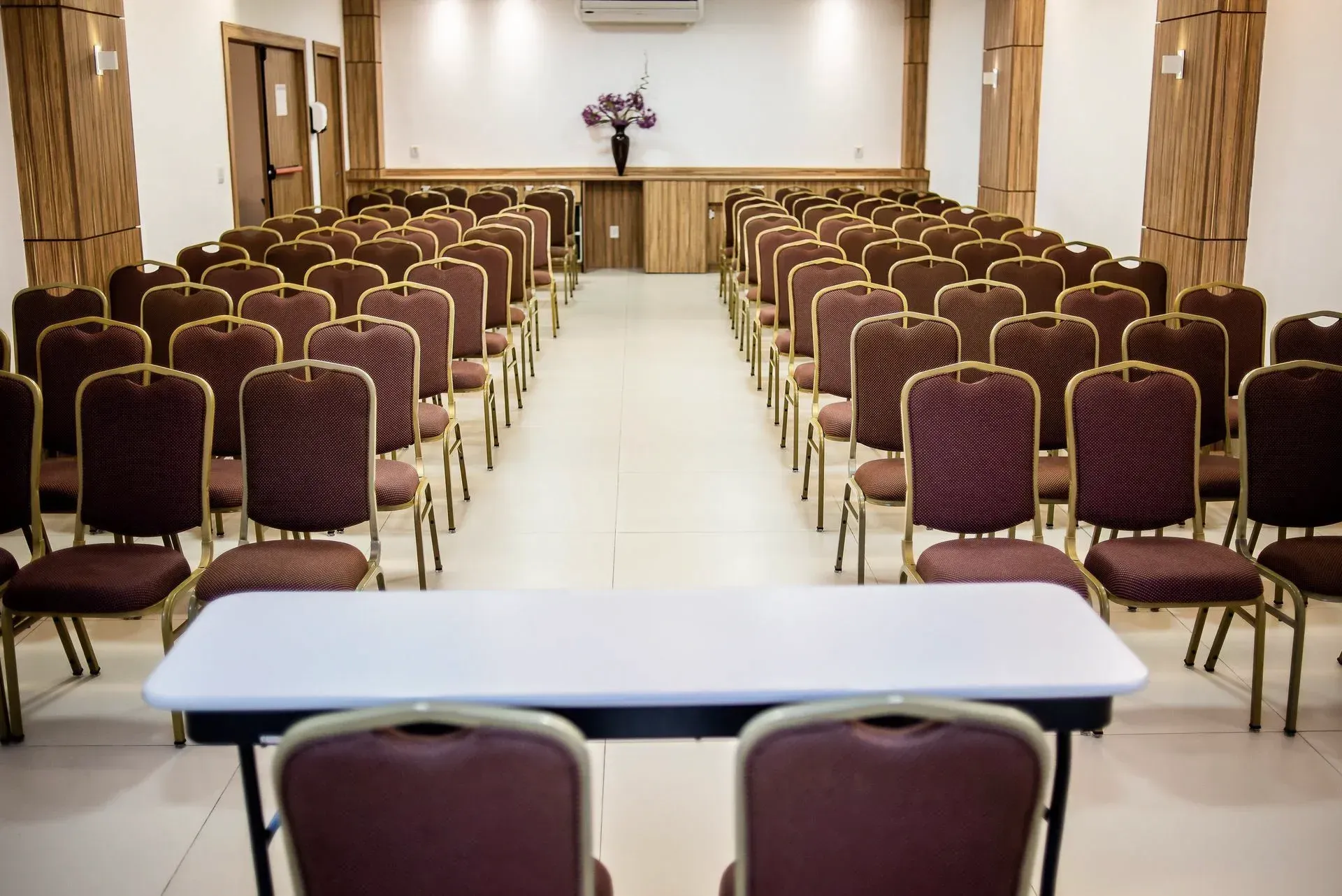 Sala de conferências vazia com fileiras de cadeiras cor de vinho e uma mesa na frente.