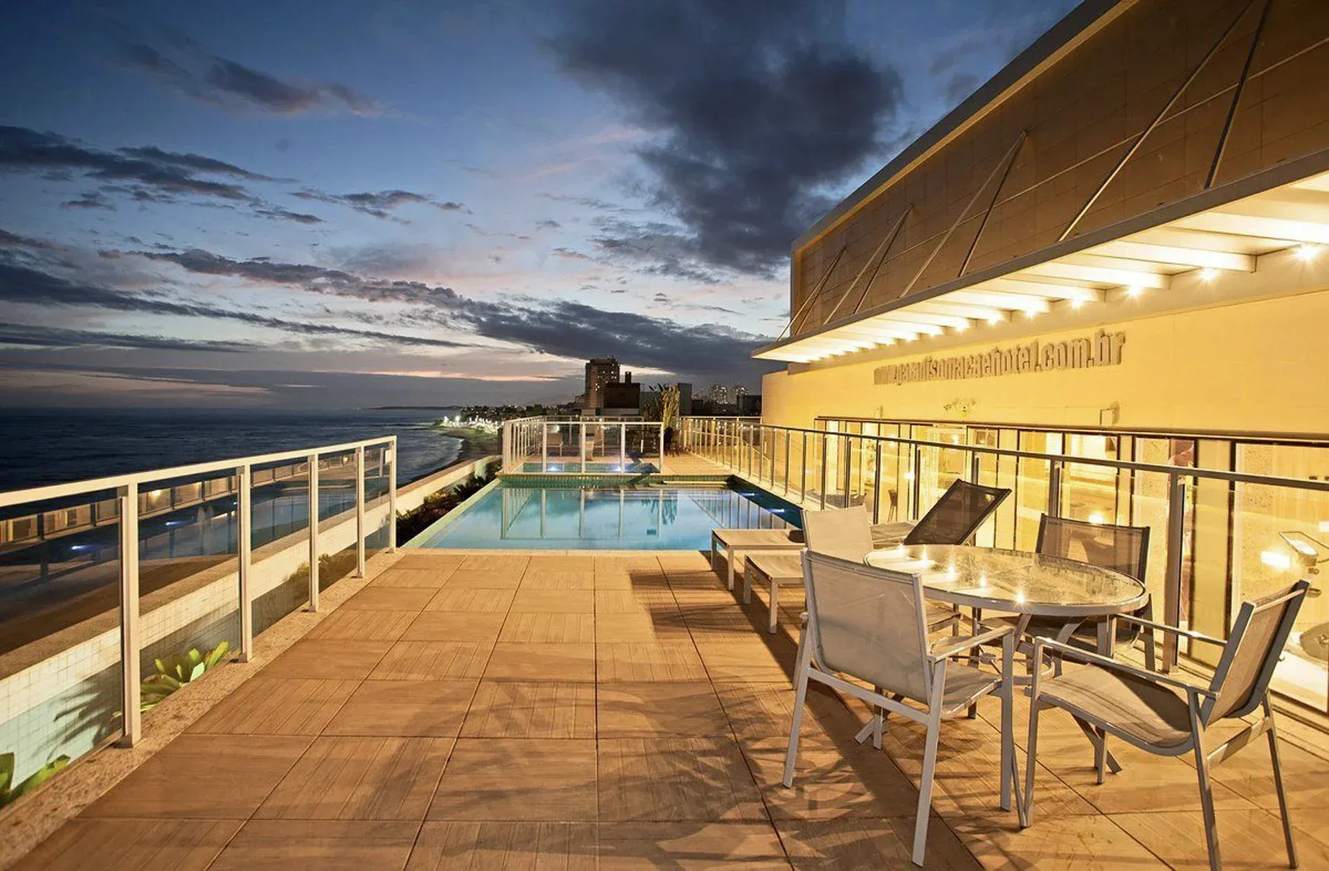 Terraço na cobertura com piscina, mesa de jantar e vista para o mar ao pôr do sol.