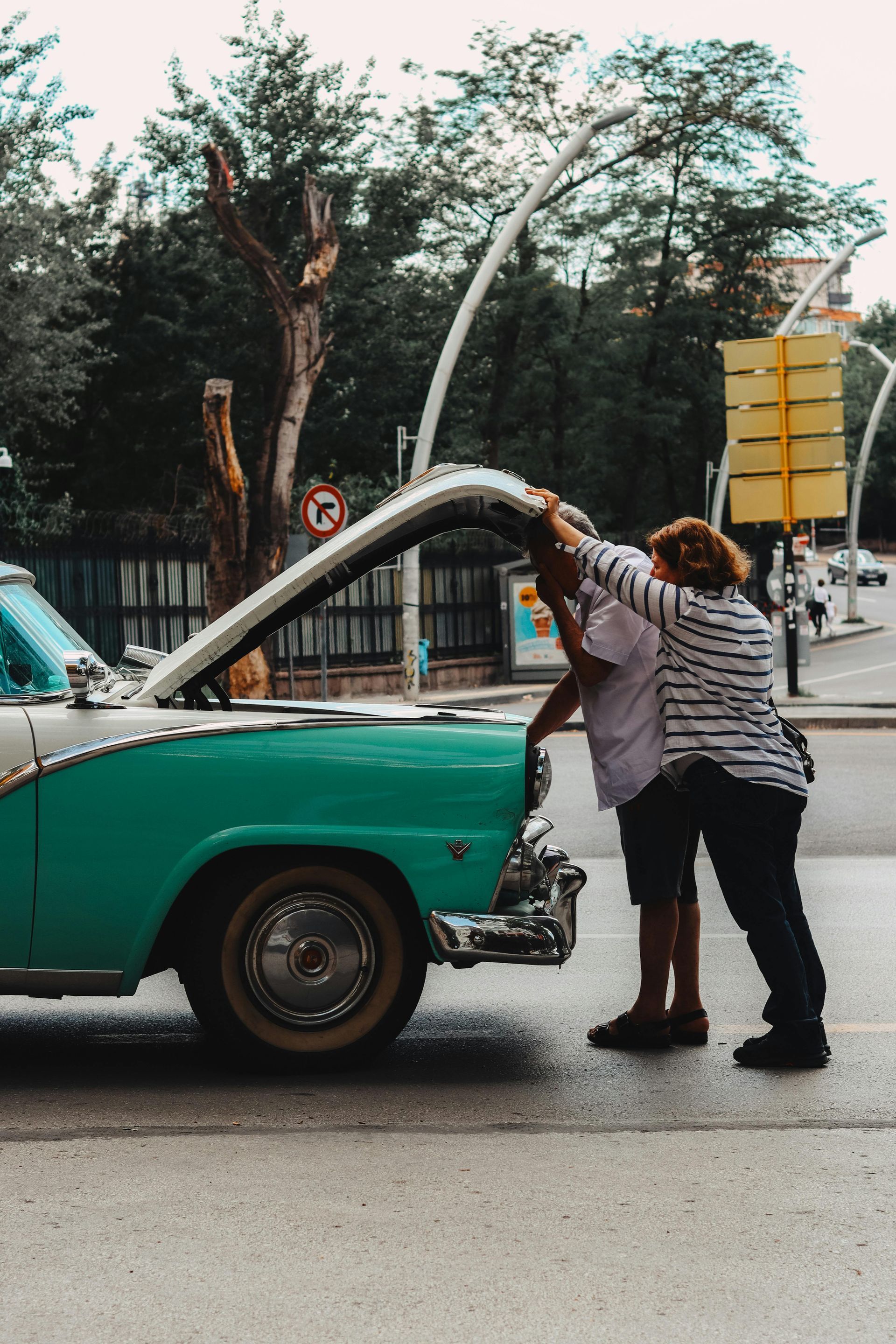 Two people refuel a turquoise vintage car on a city street.
