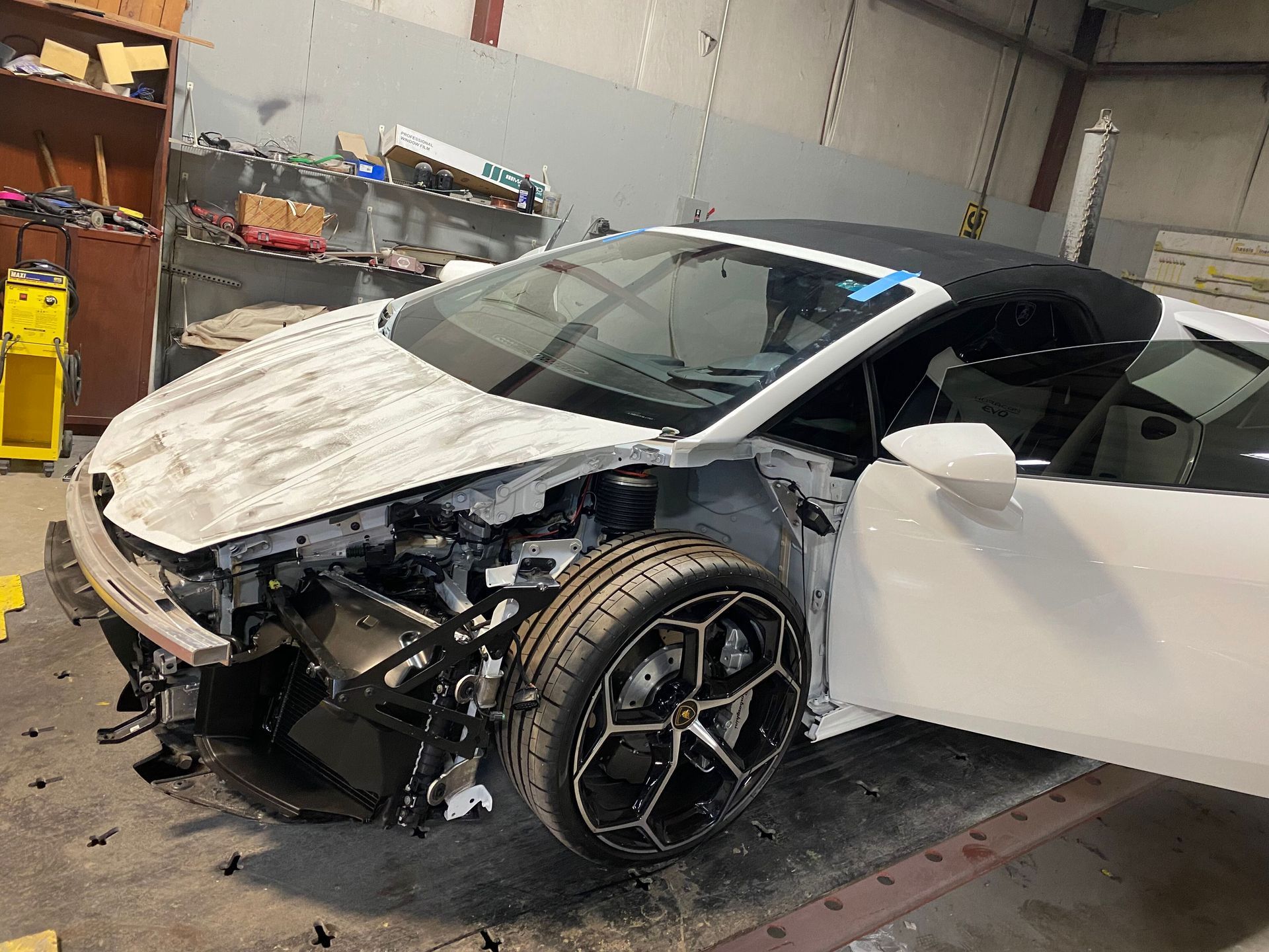 Damaged white sports car in a garage, front end crushed and right-side door open.