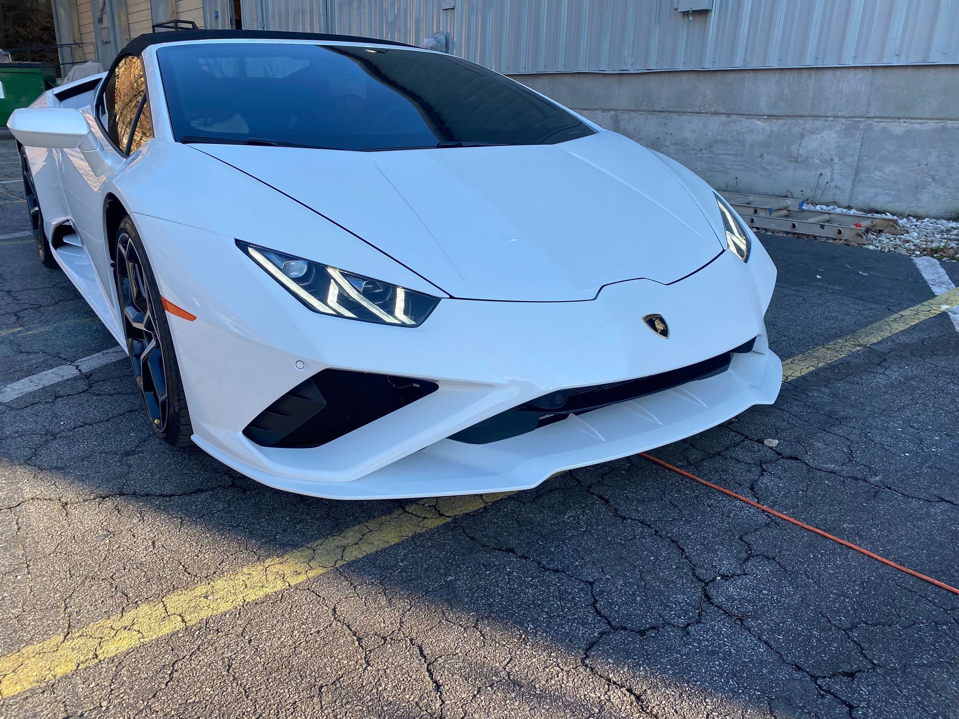 White Lamborghini parked in a lot, angled front view, with headlights on and a building wall behind it