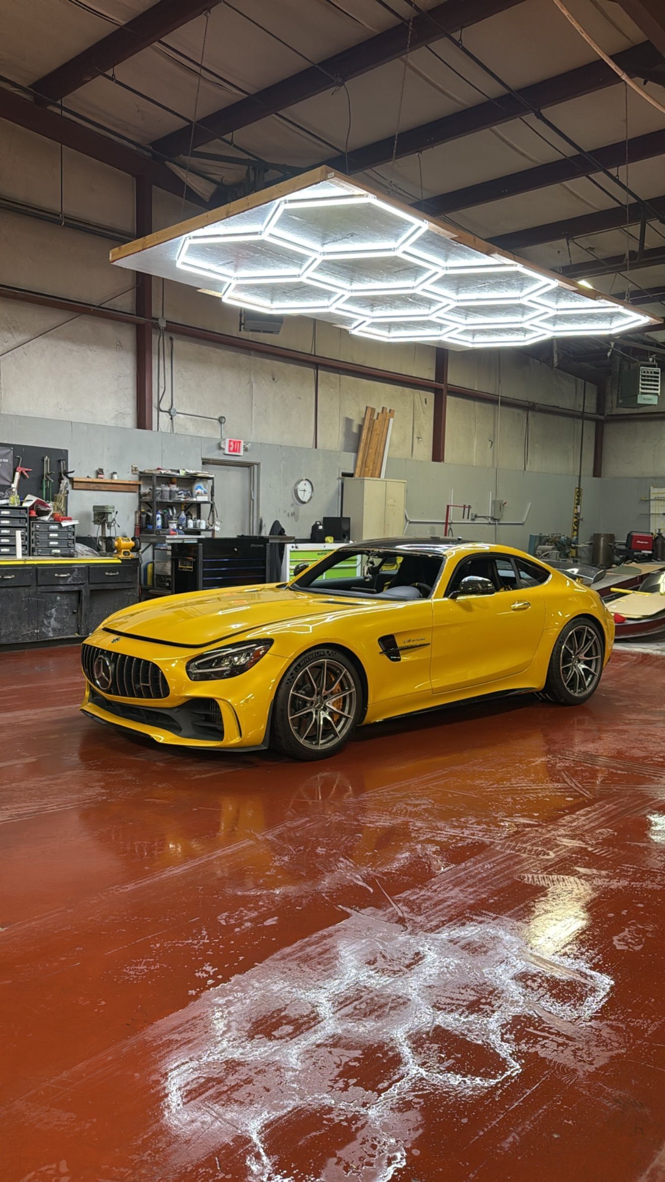 Yellow sports car in a warehouse auto shop beneath bright geometric ceiling lights.