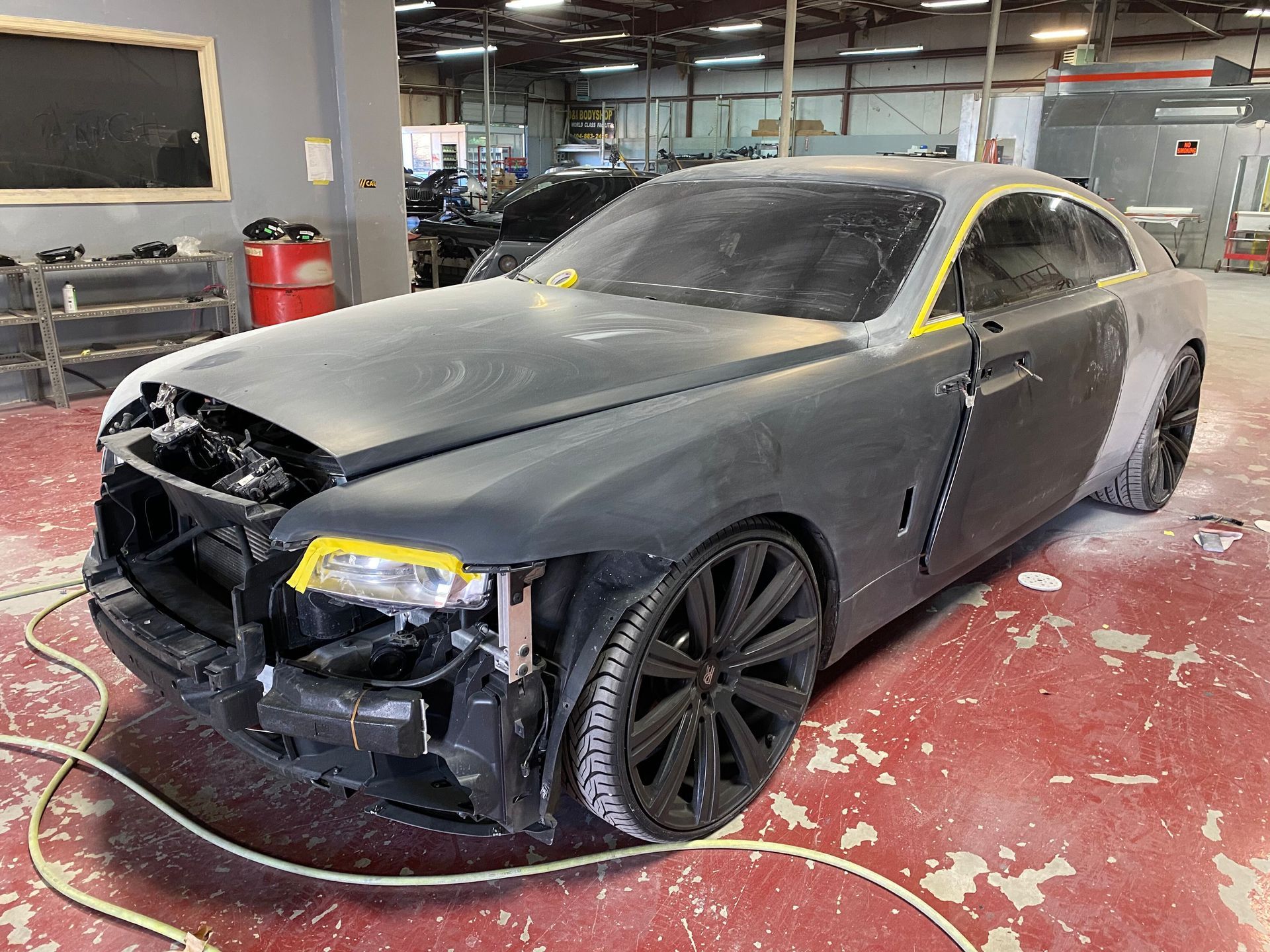 Black damaged luxury sedan in a repair shop, front bumper missing and hood taped off against a red floor.