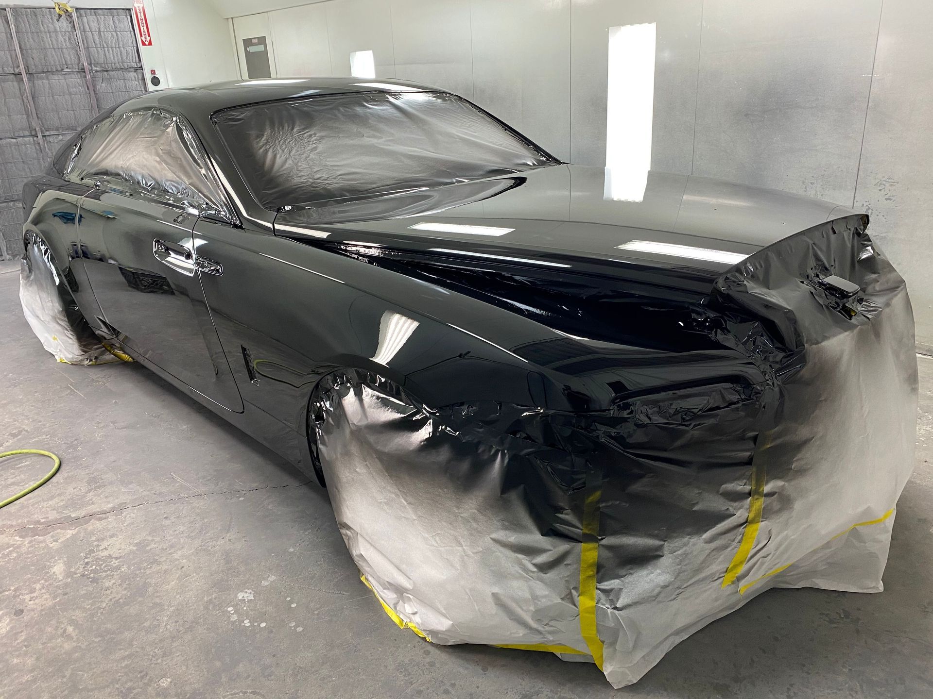 Black sedan in a repair booth, partially masked with tape and plastic for painting.