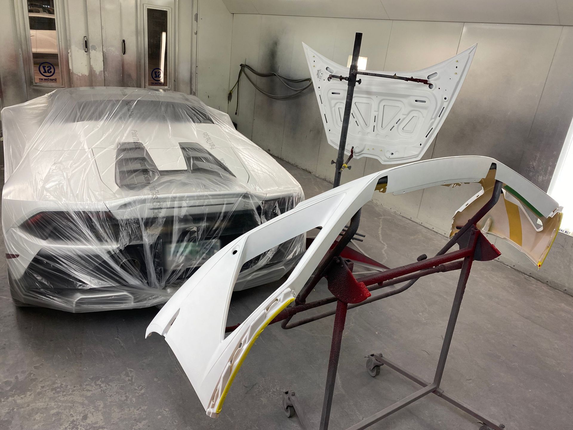 Car parts drying in a workshop: a white bumper on a red stand and another wrapped car body behind it.