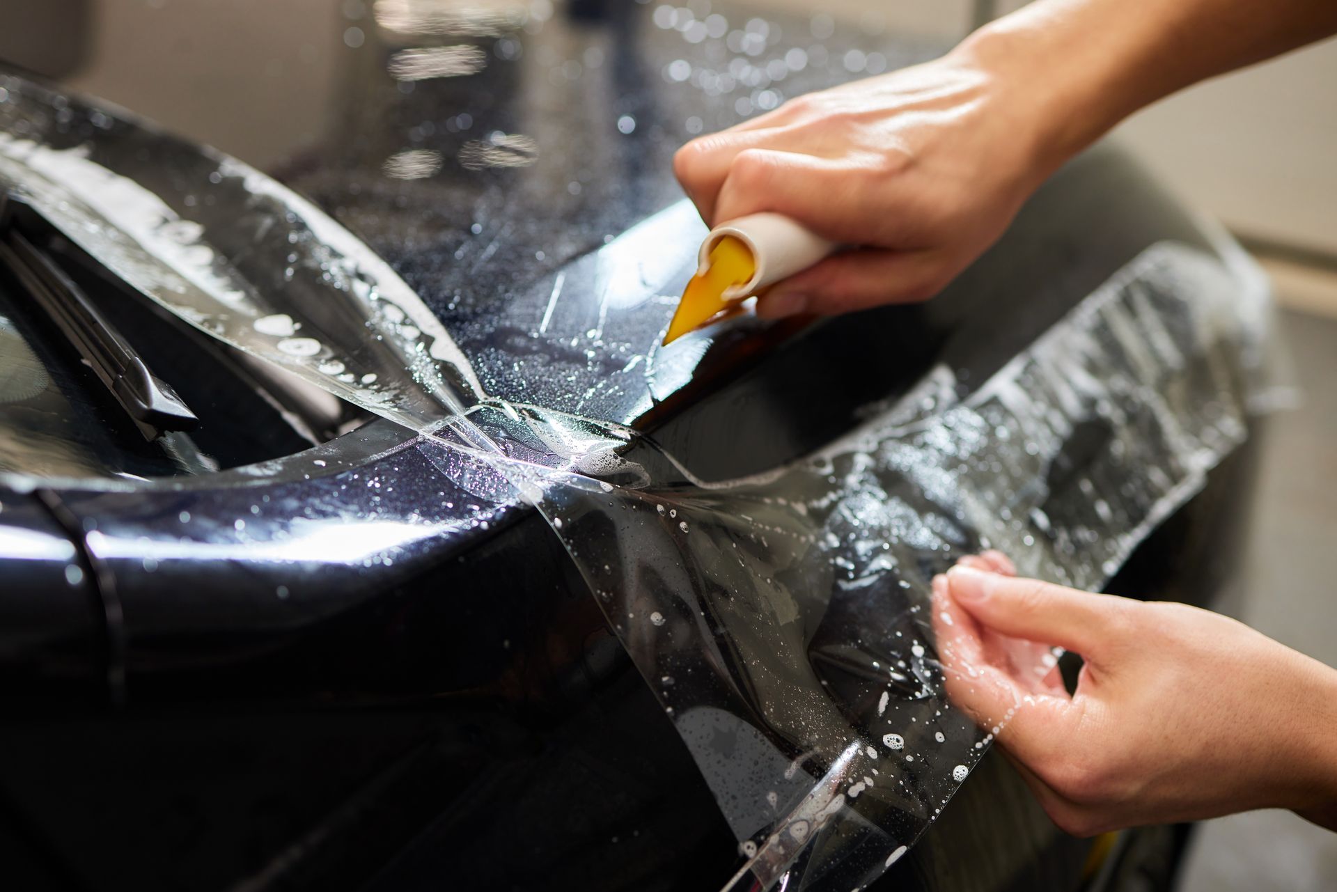 Hands applying clear film to a black car hood, smoothing out water bubbles.