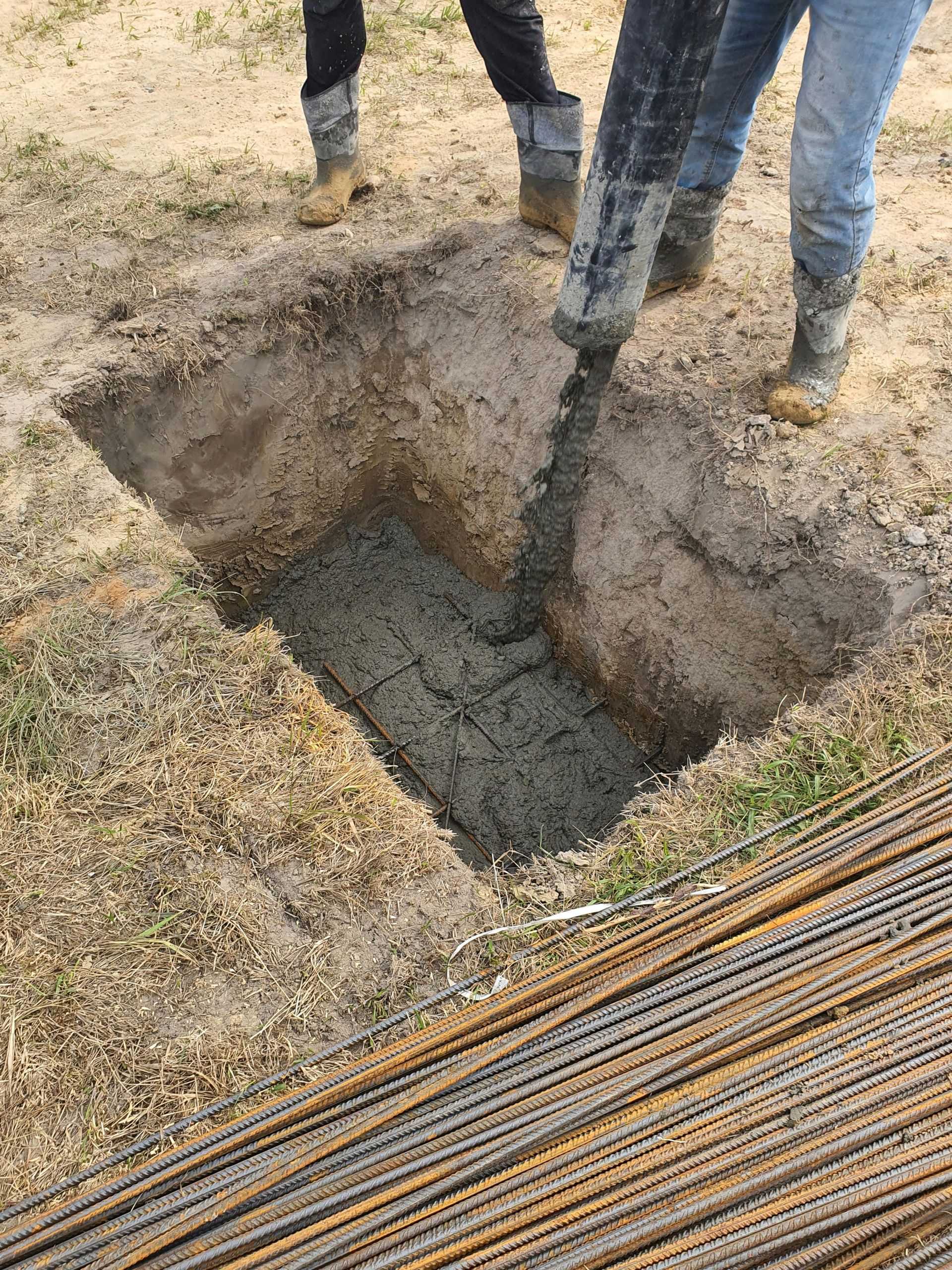 Two people pouring concrete into a rectangular foundation pit. Rebar lies beside the pit on a dirt ground.
