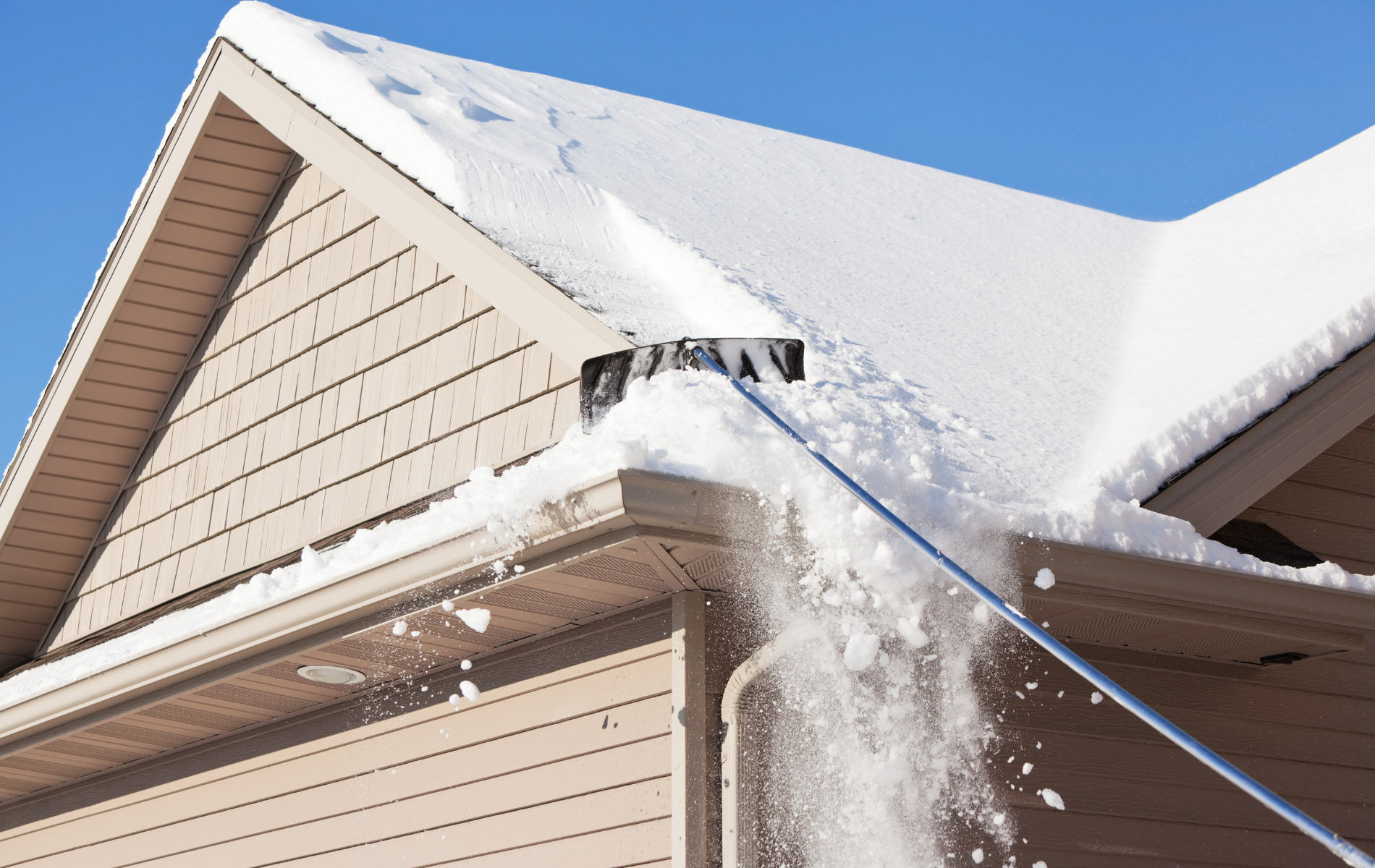 Snow being shoveled from a house roof with a long-handled snow rake on a sunny day.