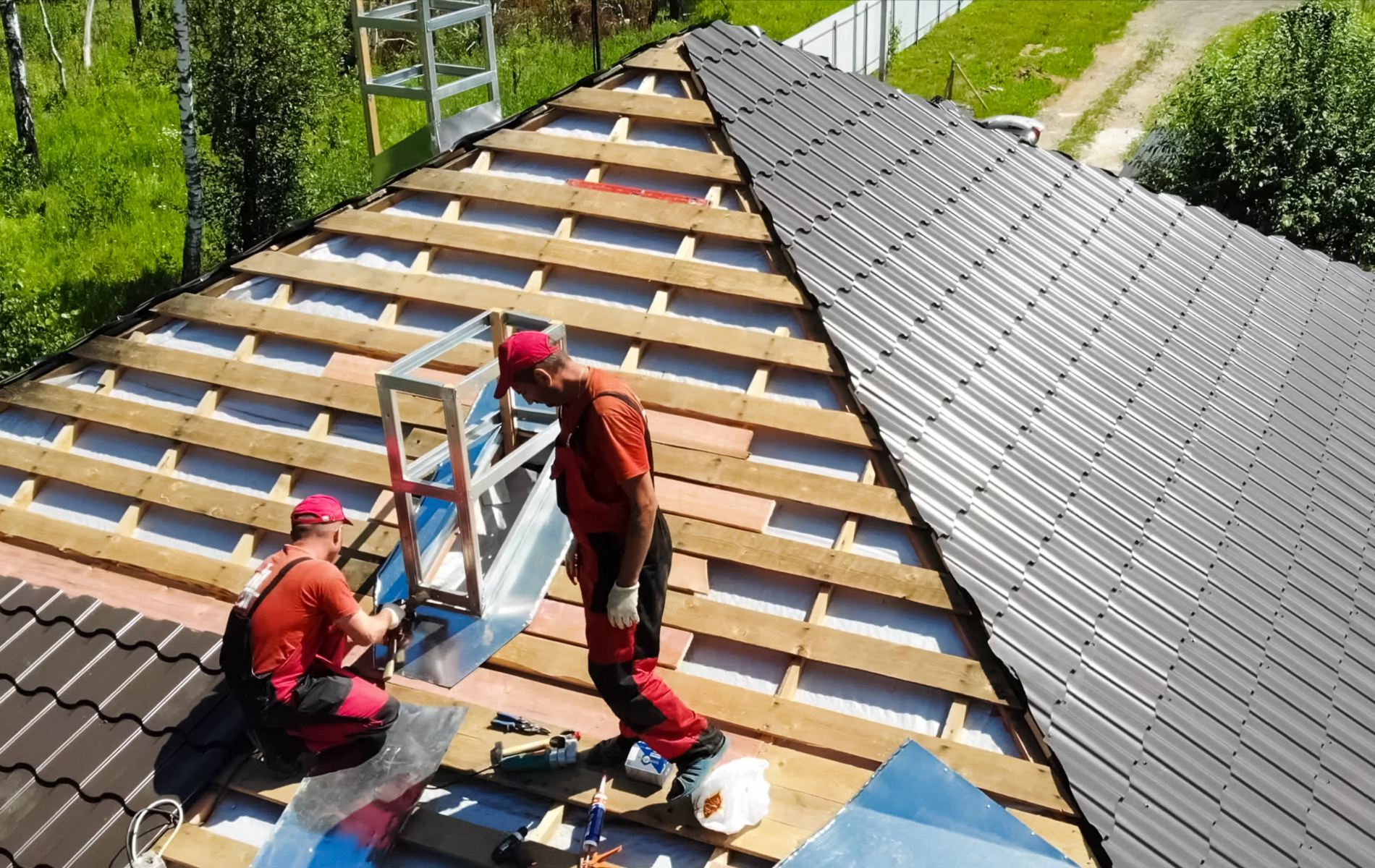 Two roofers in red work on a metal roof, securing a metal fixture.