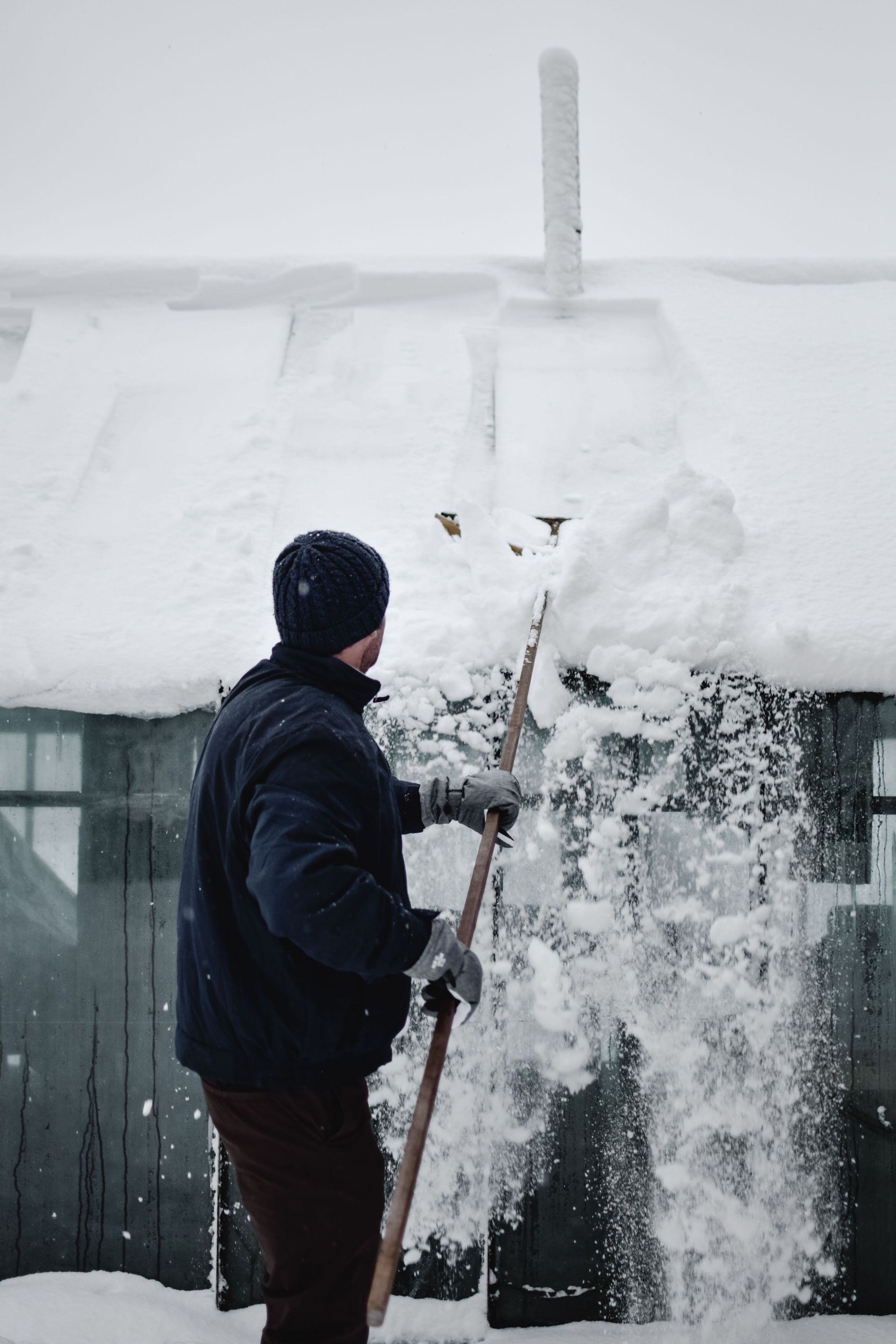 Person shoveling snow from a roof beside a snow-covered house and window