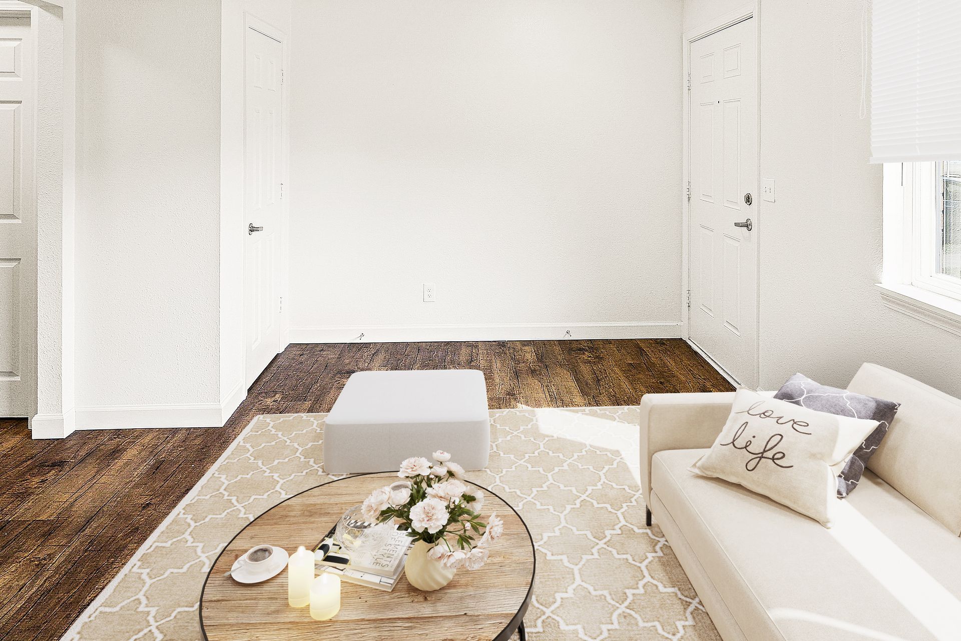 Bright, empty living room with white walls, beige rug, round coffee table, and white sofa near a window.