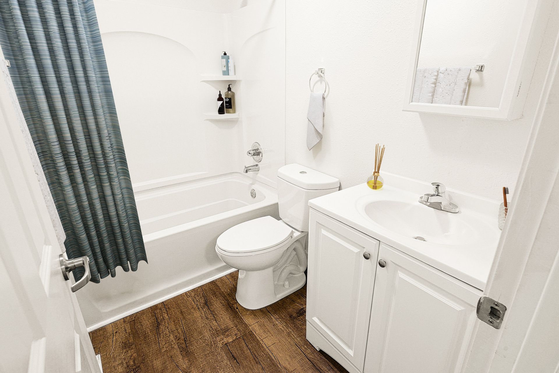 Small white bathroom with a tub, toilet, vanity sink, and gray shower curtain over wood-look flooring.
