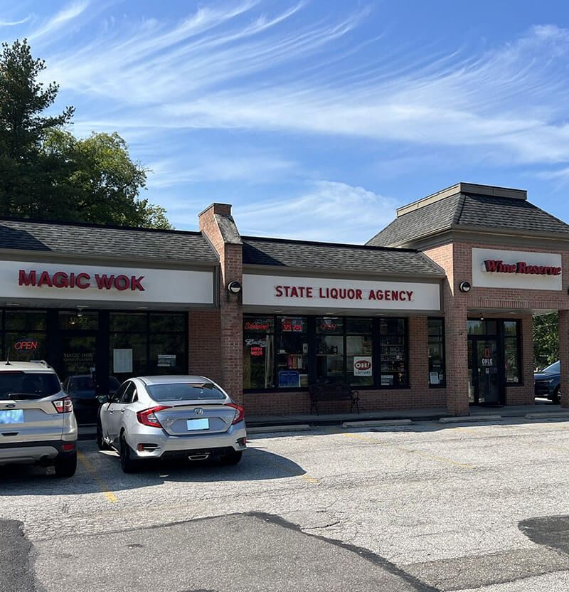 Exterior view of a strip mall with Magic Wok, State Liquor Agency, and Walgreens under a blue sky. Cars are parked in front.