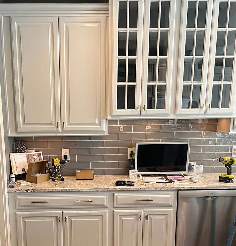 Kitchen with white cabinets, gray backsplash, and stainless steel appliances. Countertop with computer and tools.
