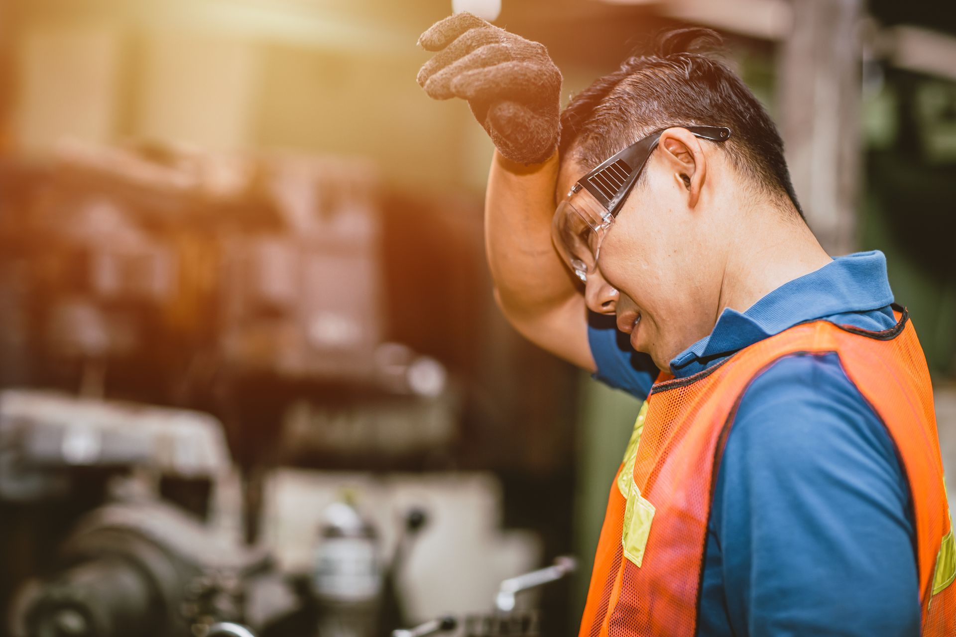 A man wearing a safety vest and glasses is wiping his forehead in a factory.