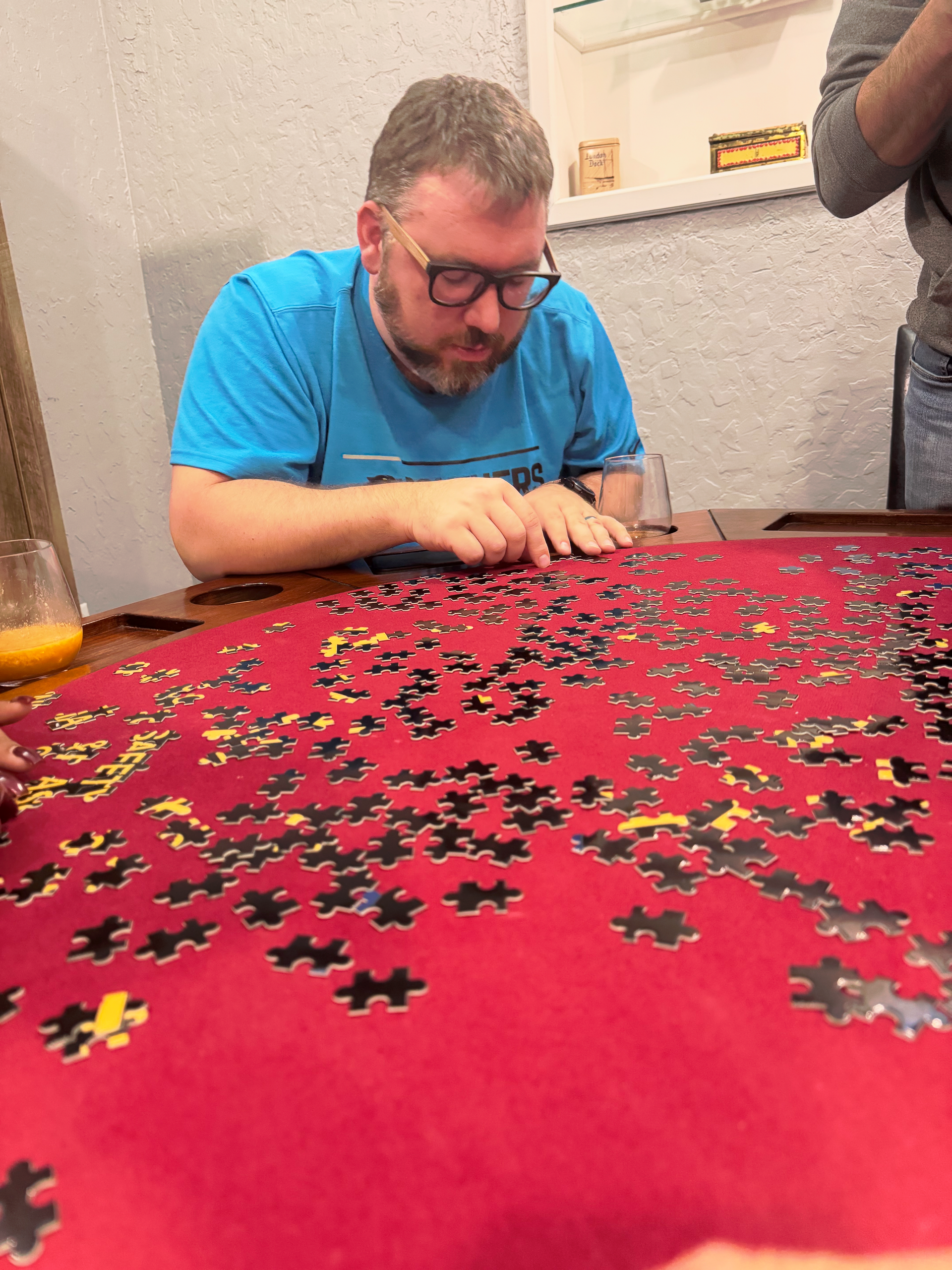 Man in blue shirt concentrating on a jigsaw puzzle spread across a red tablecloth