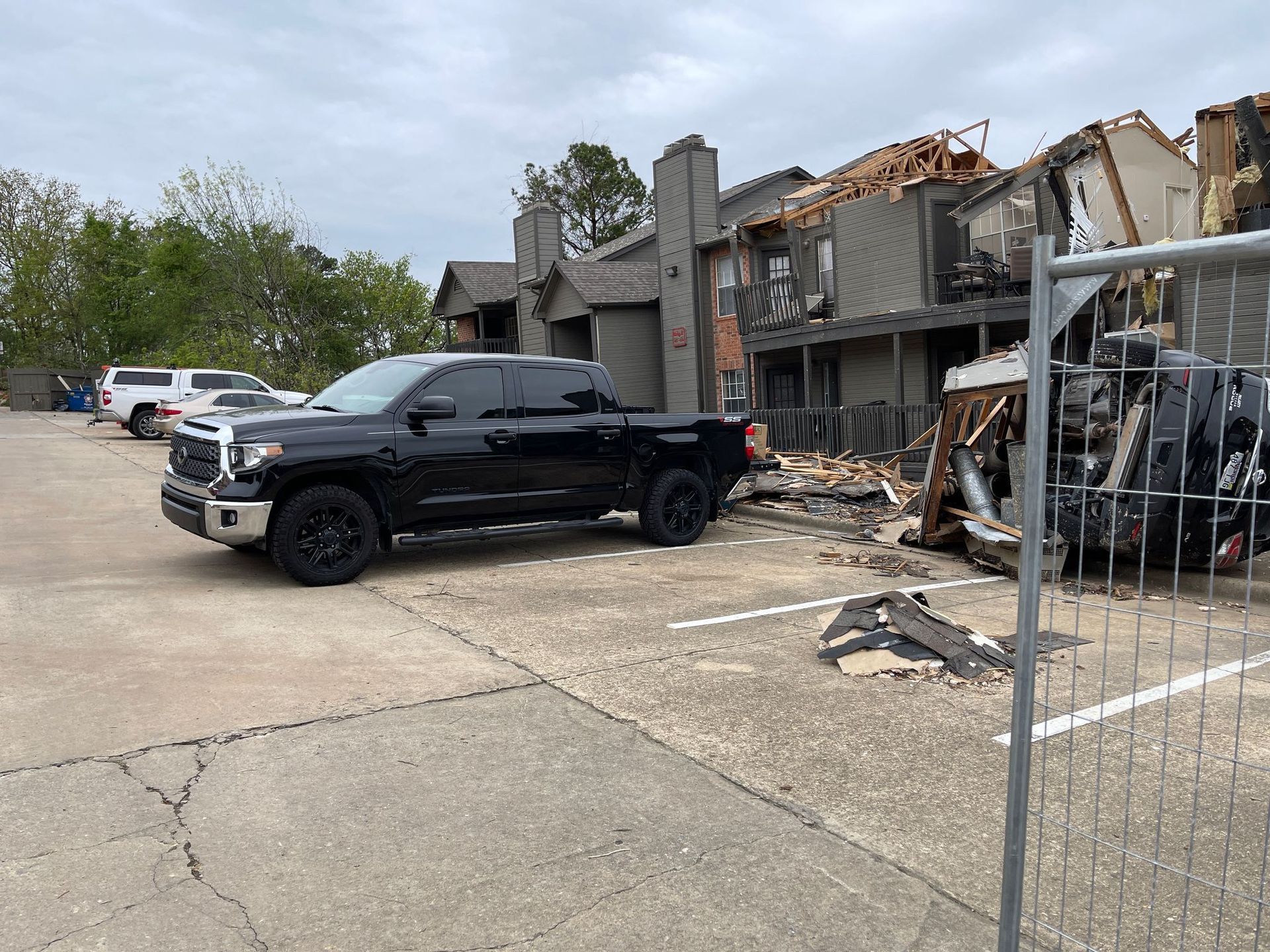 Black pickup truck parked beside a heavily damaged apartment building with debris and exposed framing.