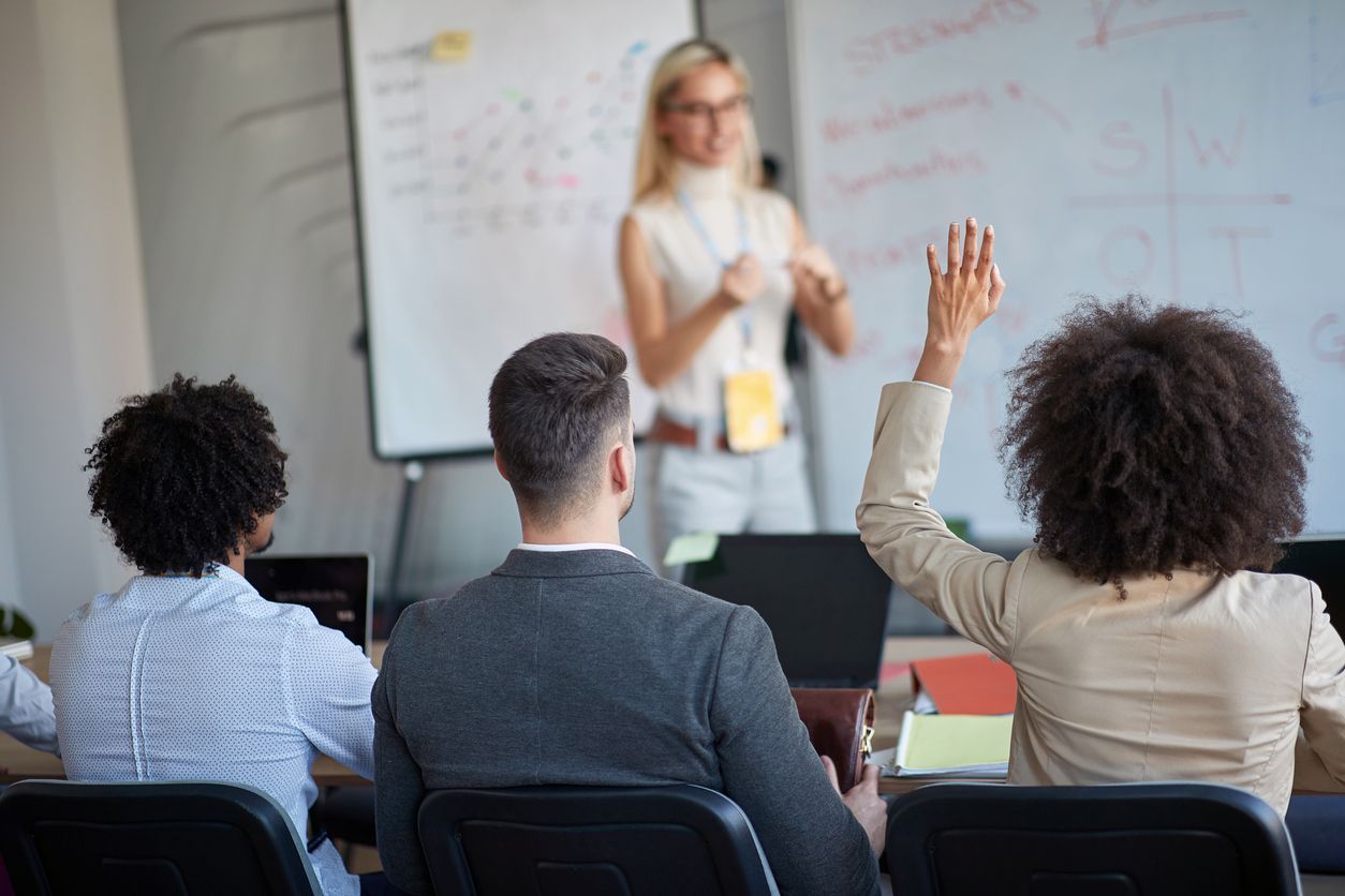 Classroom presentation with three students seated and one raising a hand toward the teacher at the whiteboard