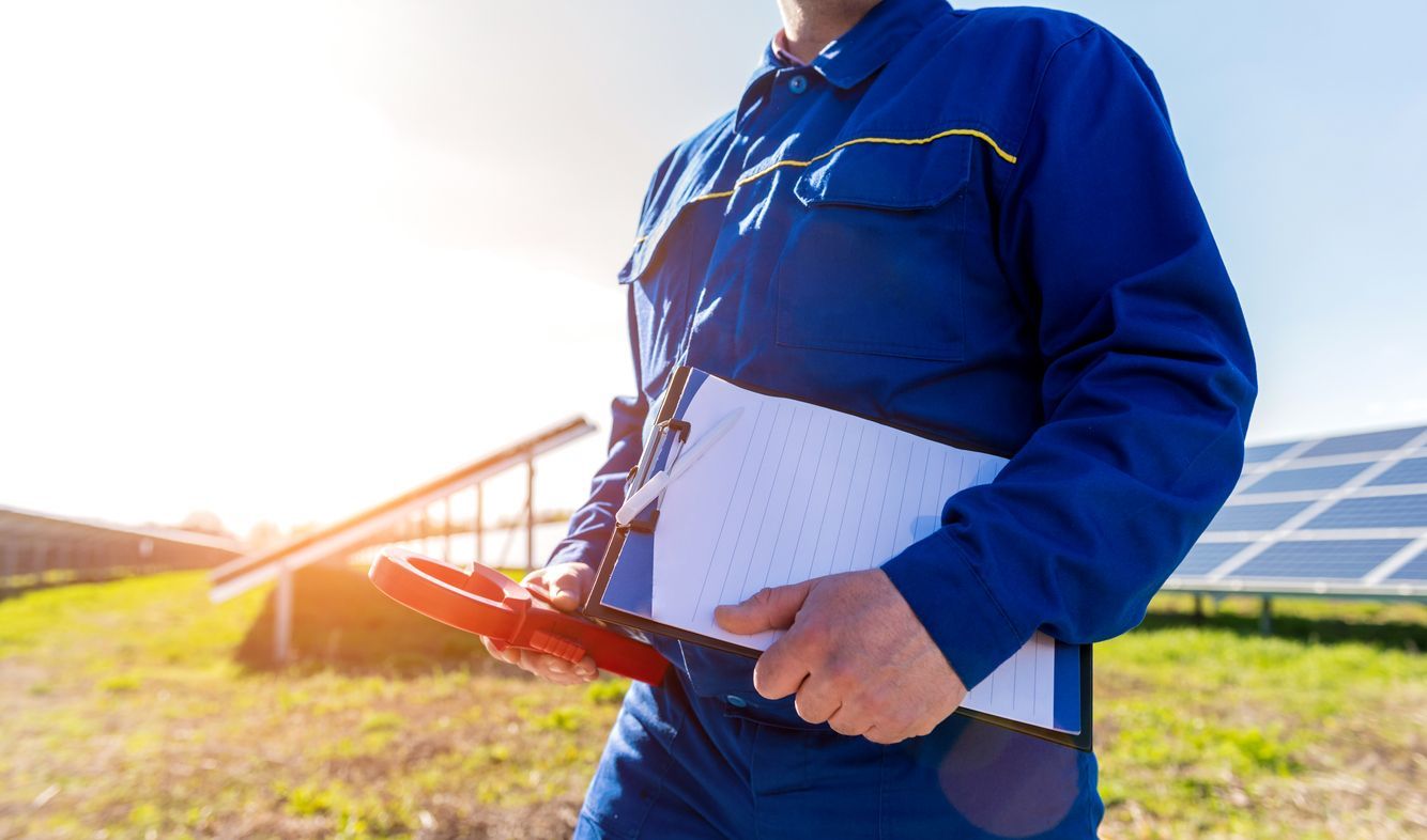 Person in blue workwear holding a tablet in a sunny field beside solar panels